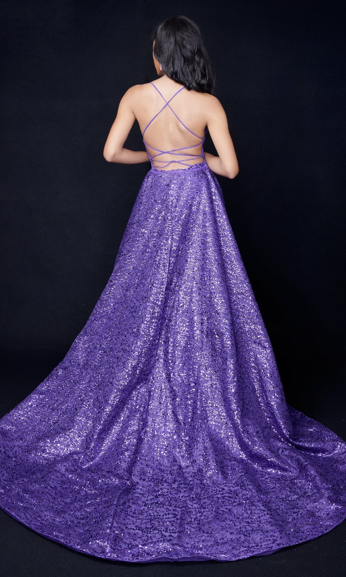 Person wearing a floor-length, backless Nina Canacci 8250 sequin gown with crisscross straps, shown from behind against a black background.