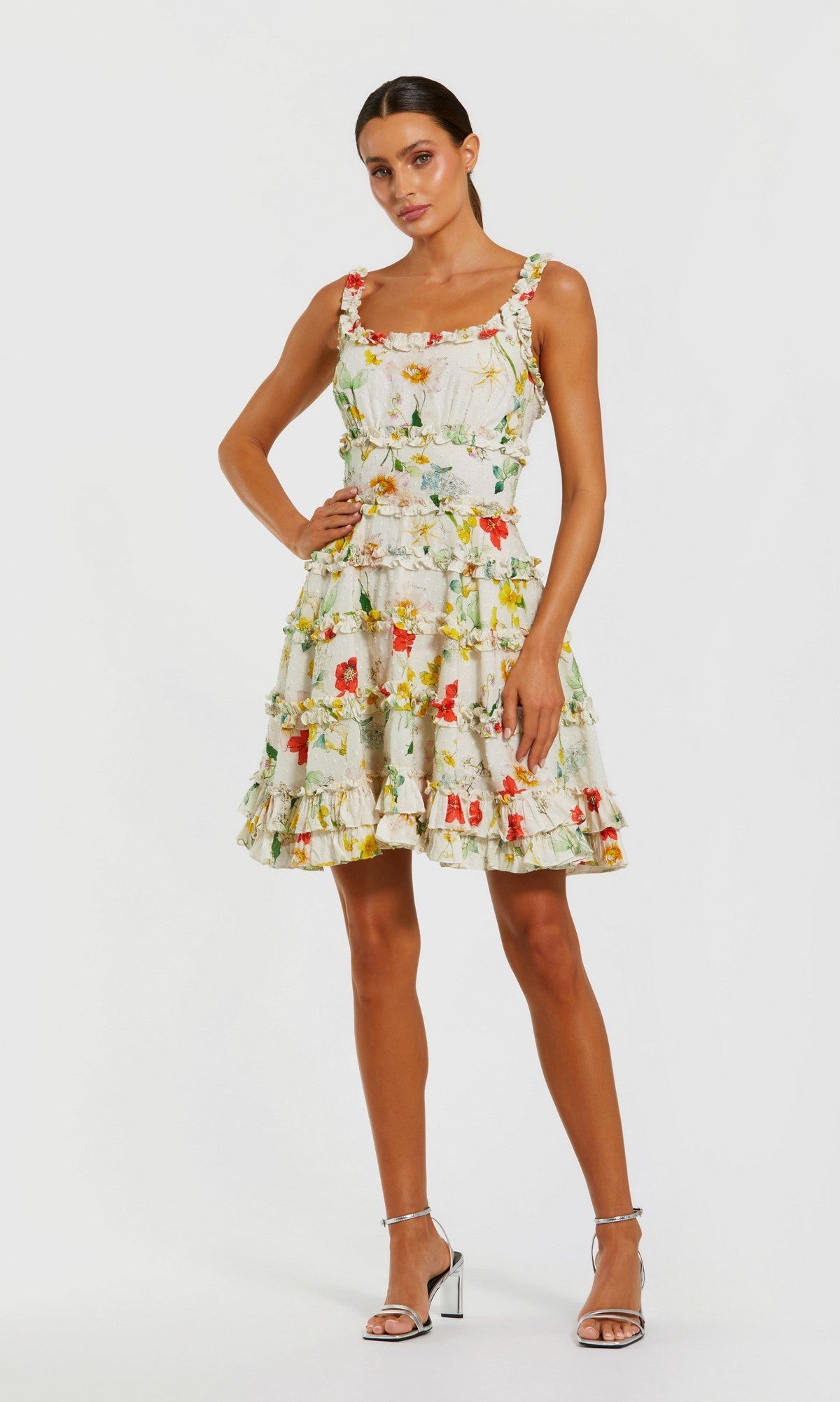 Woman in the Mac Duggal 8257 short party dress in Ivory Multi, featuring a sleeveless floral design and ruffled skirt, stands in heels against a plain white background.