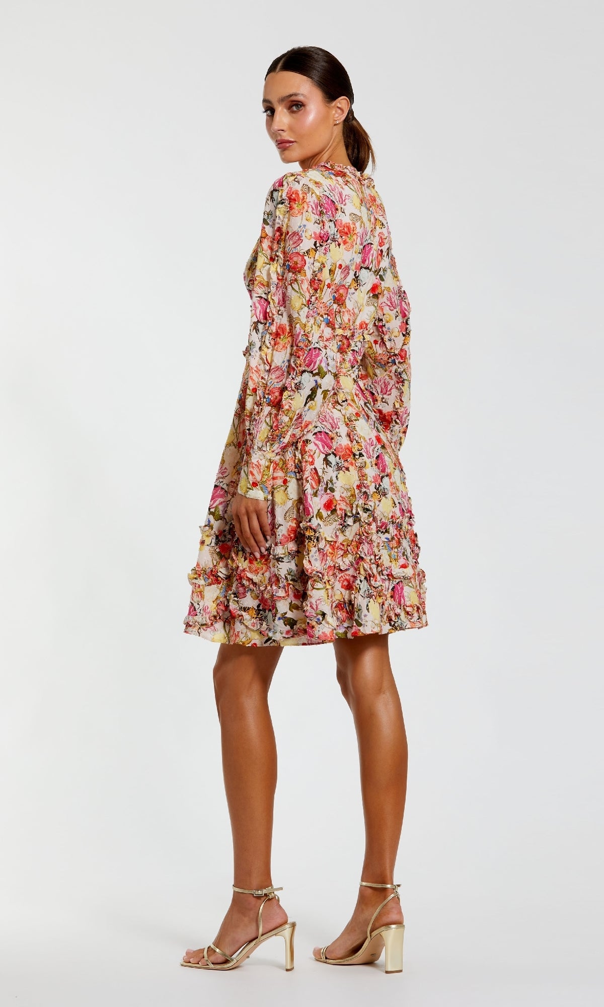 A woman models the Mac Duggal 8261 Micro-Ruffle Wedding-Guest Dress—long-sleeved, knee-length, floral-print with ruffle accents—paired with gold high heels, against a plain white background.