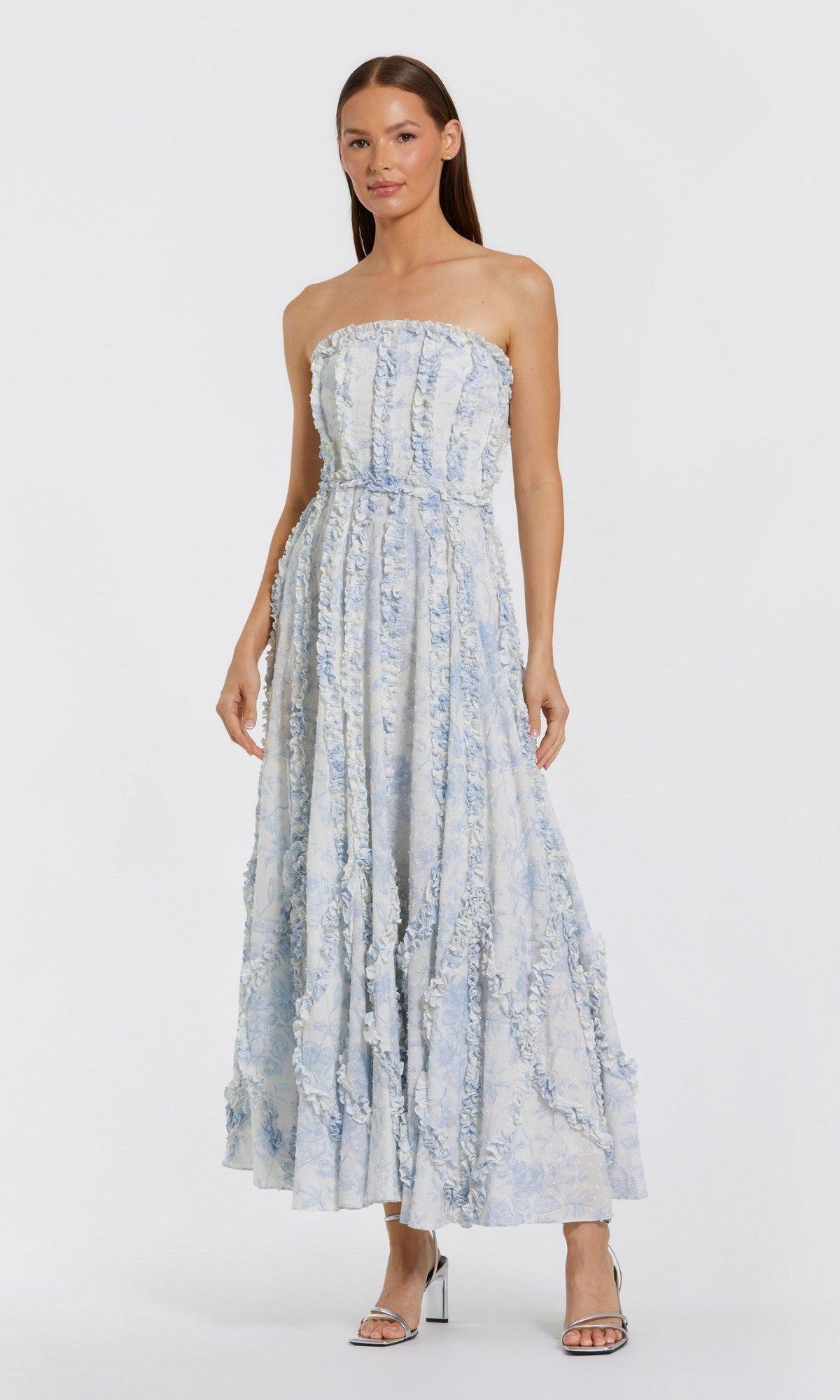 A woman stands gracefully against a plain background in the Mac Duggal 8265 Floral Wedding Guest Dress, featuring a blue multi strapless design with textured floral patterns, tea-length hem, and silver high-heeled sandals.