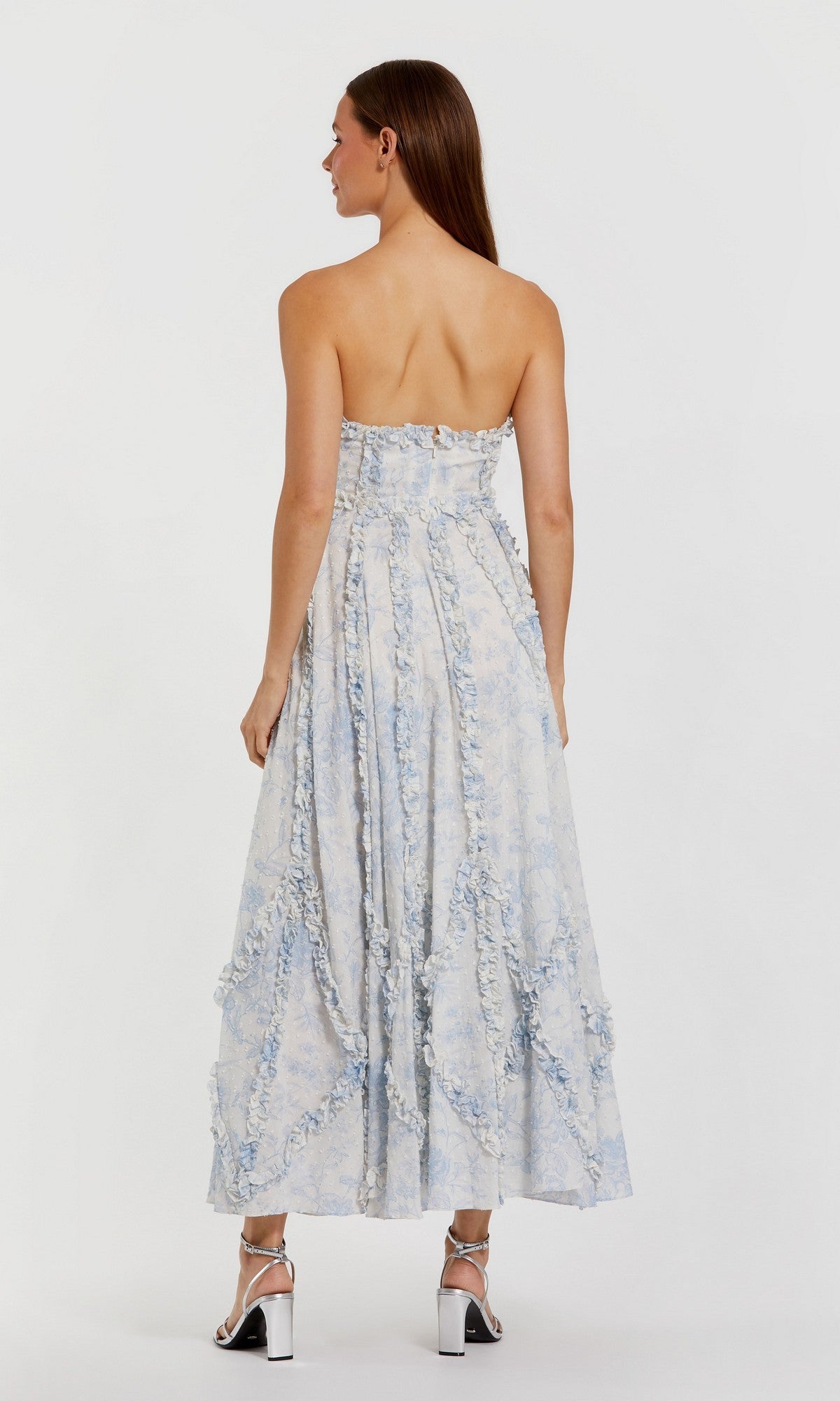A woman with long brown hair stands with her back to the camera, wearing the Mac Duggal 8265 Floral Wedding Guest Dress in light blue and white cotton, paired with silver high-heeled sandals.