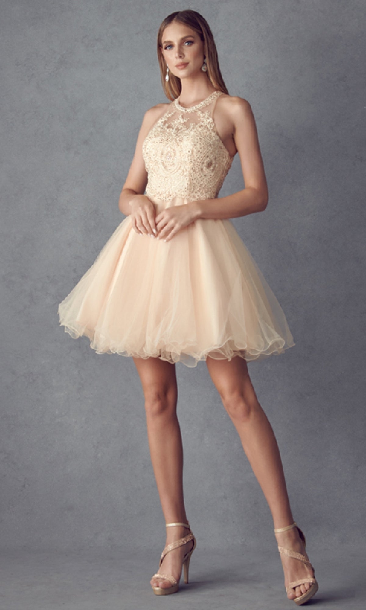 Woman models the Short Hoco Dress: Juliet 826—a sleeveless, beige lace and tulle mini dress with an a-line skirt and fitted bodice—paired with strappy heels against a gray backdrop for a chic homecoming look.