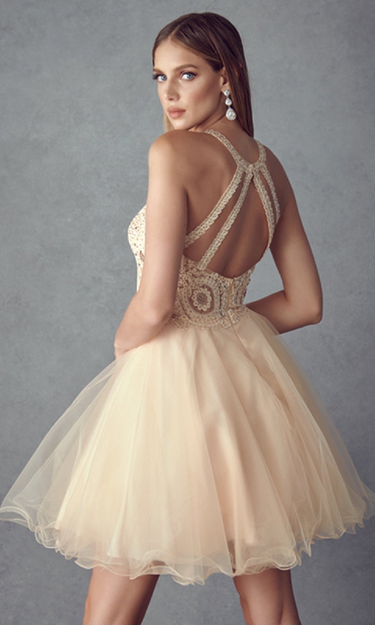 Woman wearing the Juliet 826 Short Hoco Dress, a light beige sleeveless babydoll style with a lace bodice, open strappy back, and A-line tulle skirt, standing against a plain grey background.