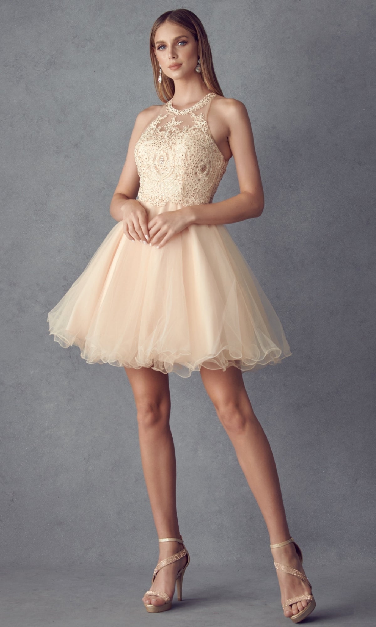 A woman poses against a gray background wearing the Juliet 826 short Hoco dress—sleeveless, beige lace with an A-line tulle skirt—paired with nude high-heeled sandals.