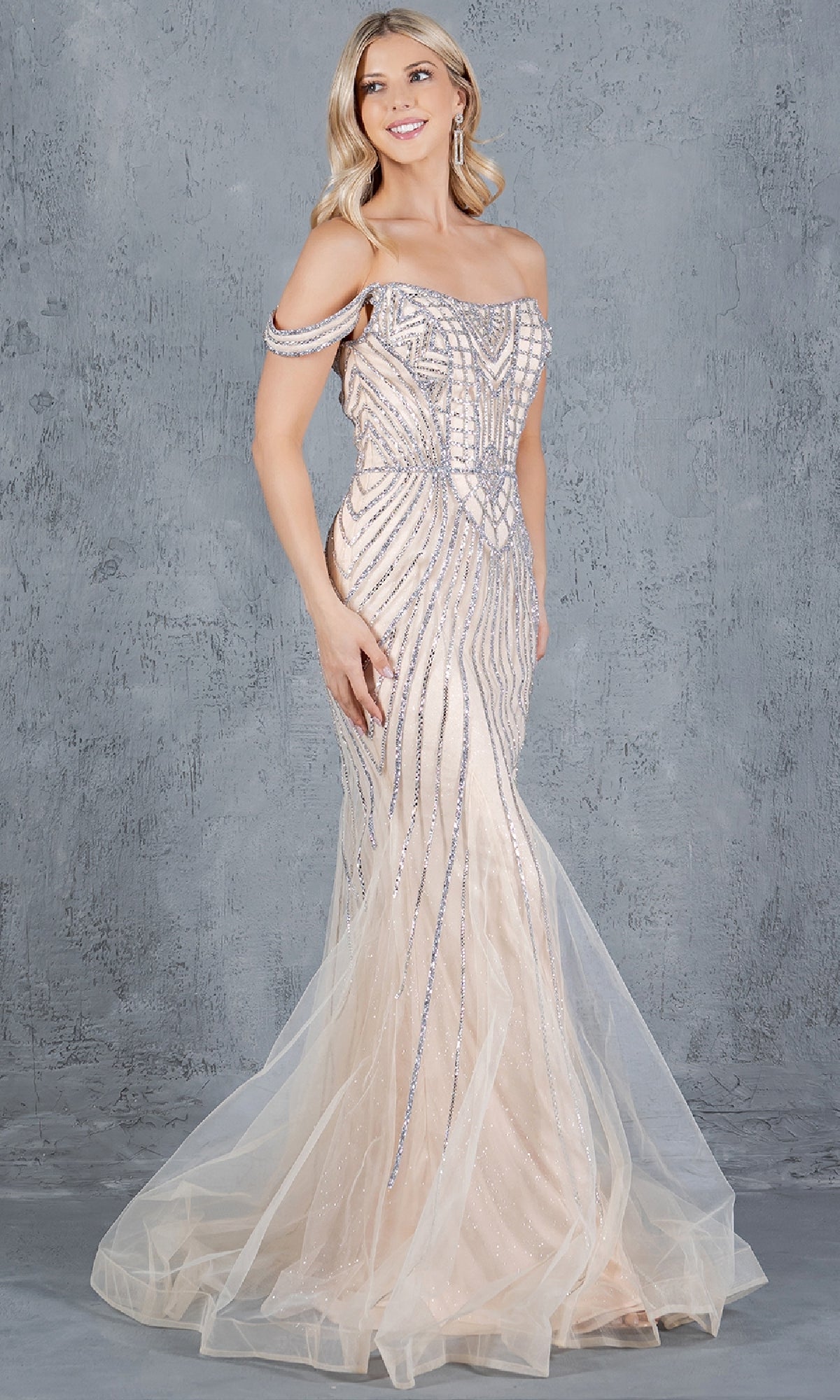 A woman models the Long Beaded Prom Dress: Cinderella Couture 8271J, an elegant light beige off-the-shoulder gown with silver embellishments, standing against a textured gray background.