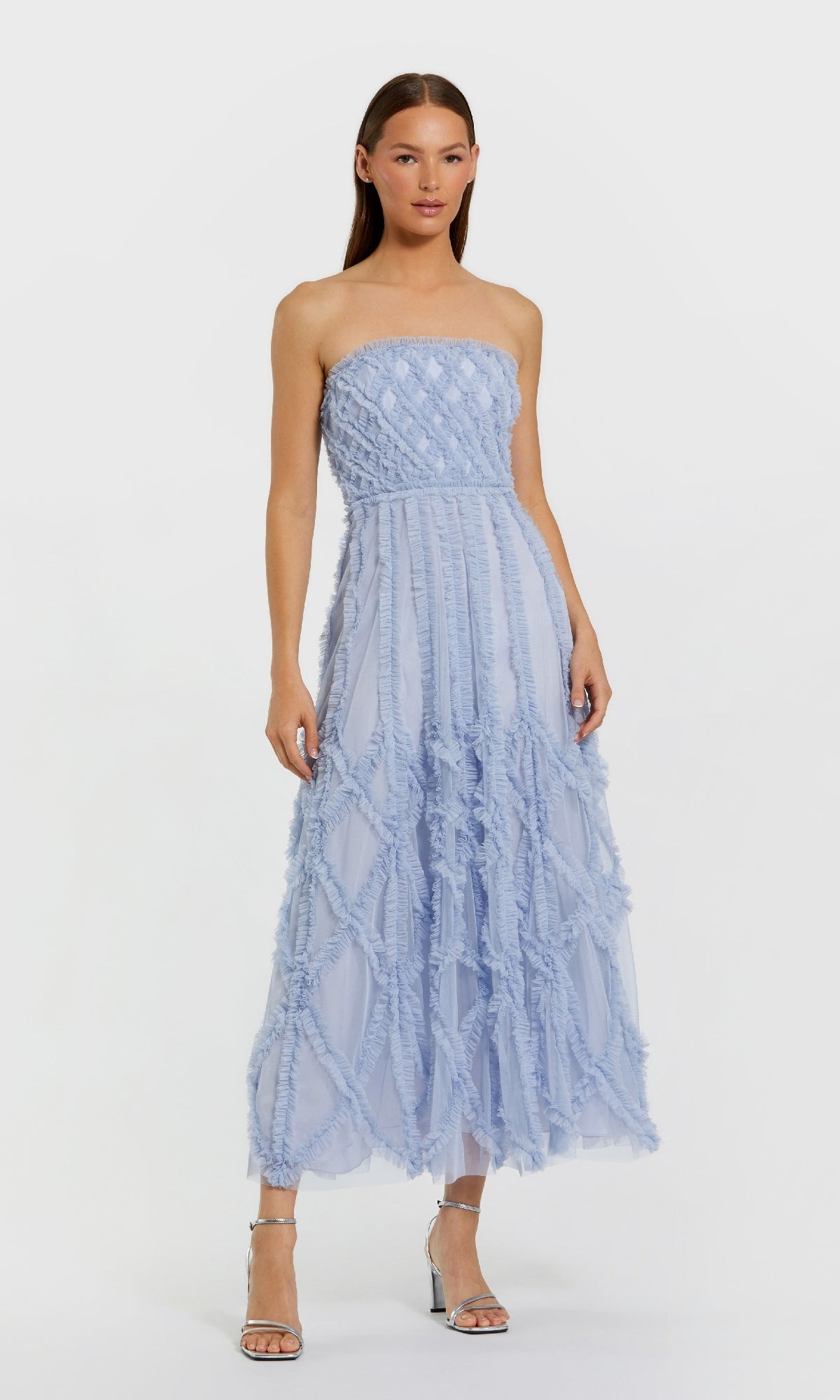 A woman stands against a white background wearing the Periwinkle Wedding Guest Dress: Mac Duggal 8277, a strapless light blue dress with textured crisscross detailing, paired with silver high-heeled sandals.