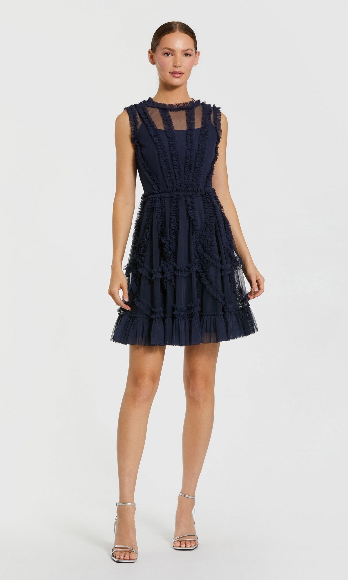 A woman stands against a white background wearing the Mac Duggal 8289 short wedding guest dress in indigo, a sleeveless fit and flare style with sheer paneling and ruffle details, paired with silver high-heeled sandals.