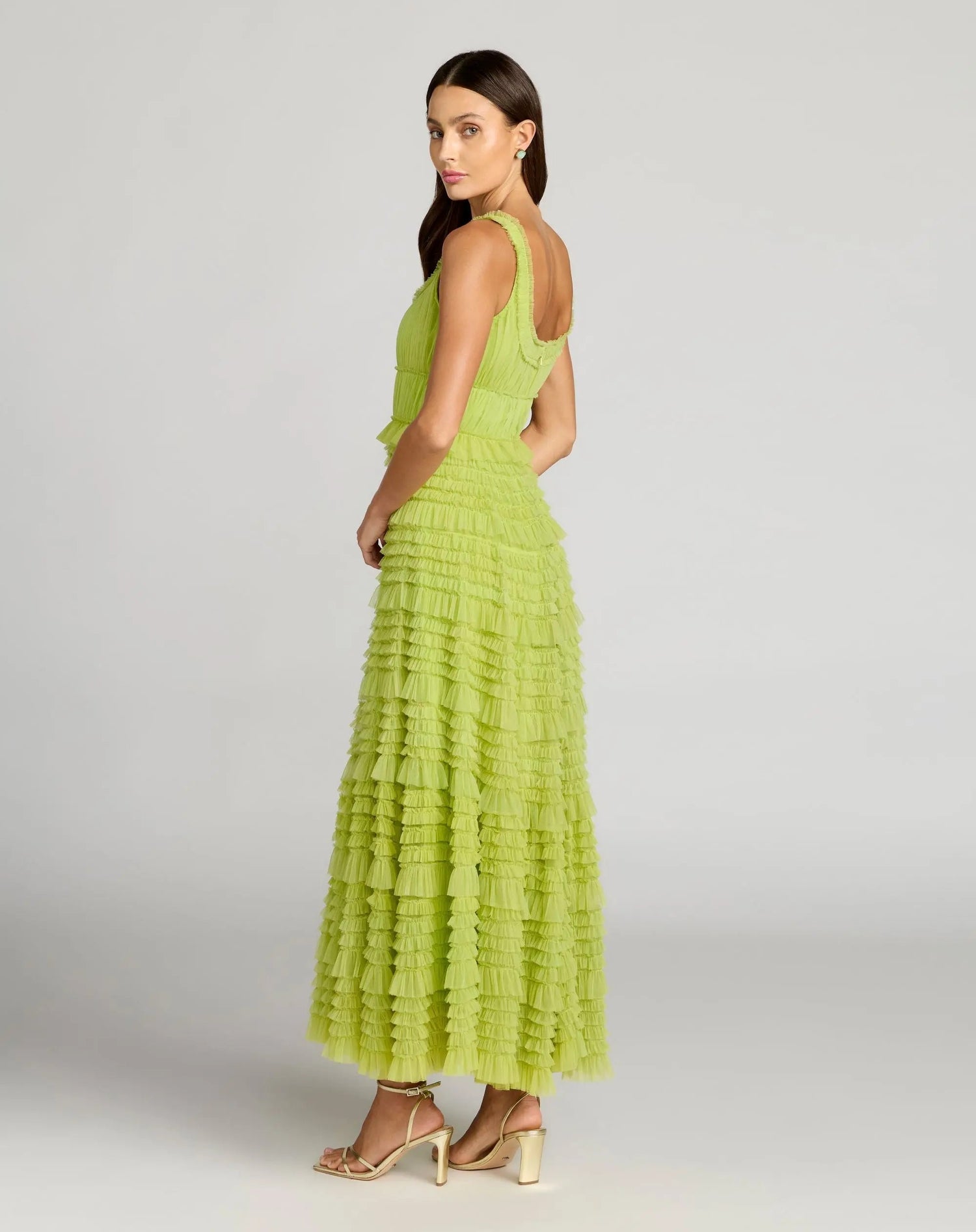 A woman stands in profile against a plain gray background, wearing the Mac Duggal 8292 tea-length party dress—a sleeveless lime green gown featuring ruffle detailing and an elegant A-line tiered skirt—paired with beige open-toe heels.