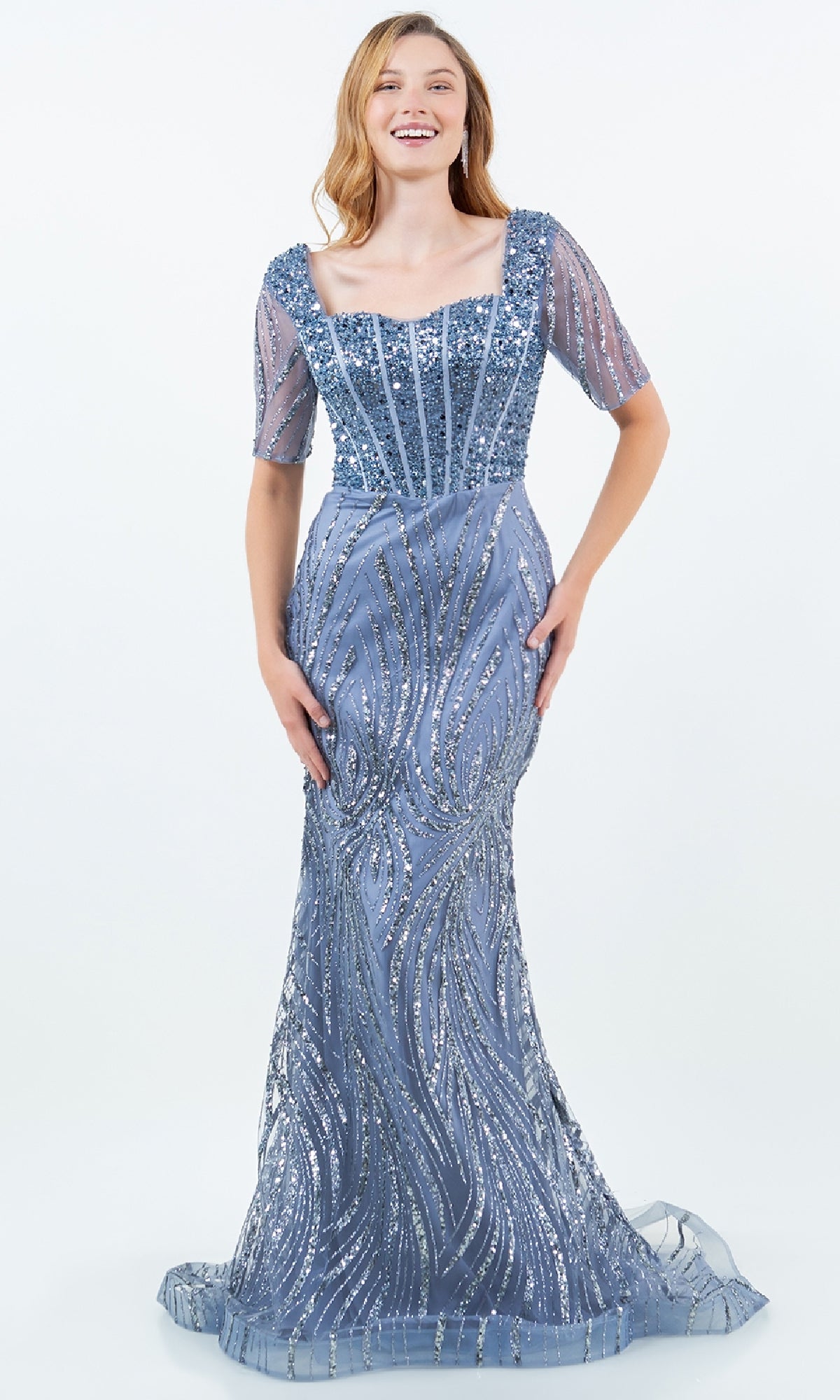 A young woman models the Cinderella Couture 8293M, a floor-length blue sequin mermaid gown with bead accents, short sheer sleeves, and a square neckline, standing against a plain white background.