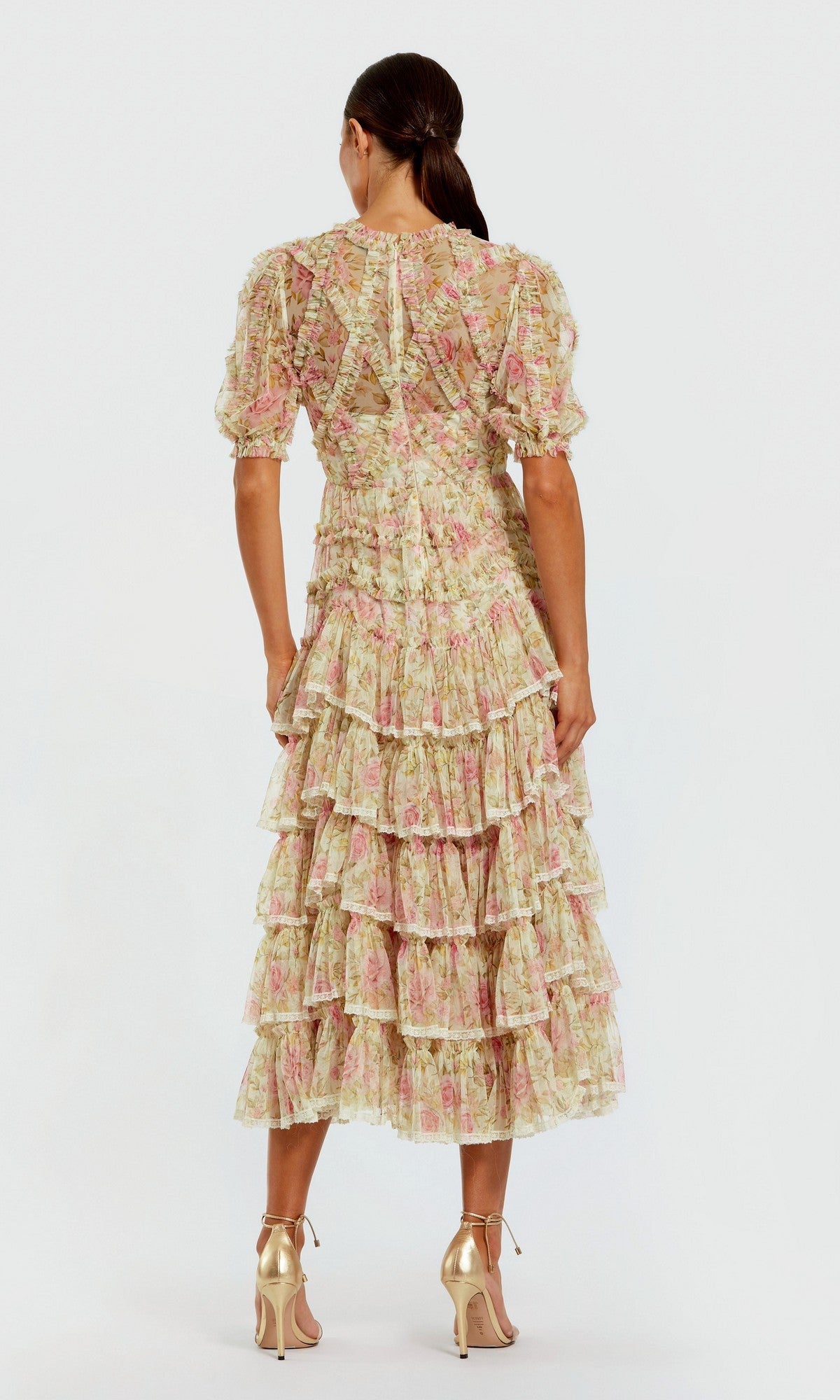 A woman stands facing away, wearing the Midi Wedding Guest Dress: Mac Duggal 8295, a pink and cream floral mesh midi dress with ruffle details, puff sleeves, and gold high-heeled sandals.