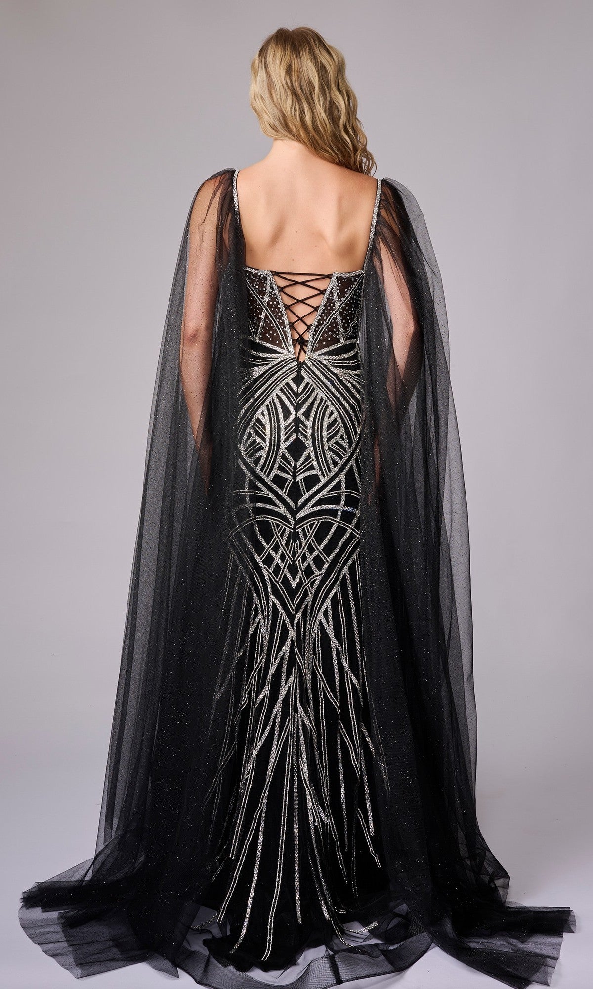 A woman in the Nina Canacci 8305 long formal dress, featuring silver geometric embellishments and a sheer black cape, stands facing away to reveal the lace-up back detail of her gown.