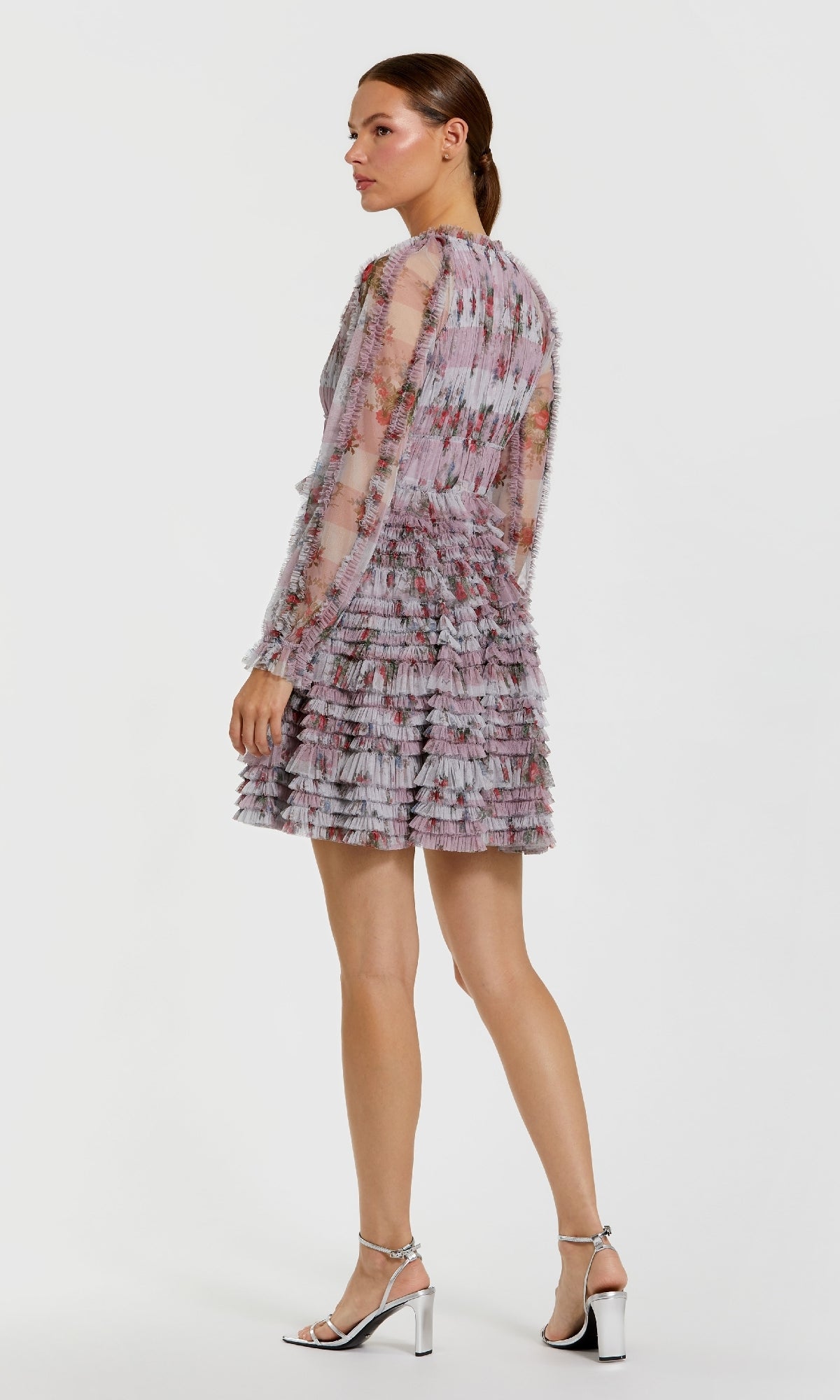 A woman stands in profile wearing the Mac Duggal 8328 Short Wedding Guest Dress, a sheer, long-sleeved mini with ruffles and a floral-inspired pattern, paired with silver high-heeled sandals against a plain white background.
