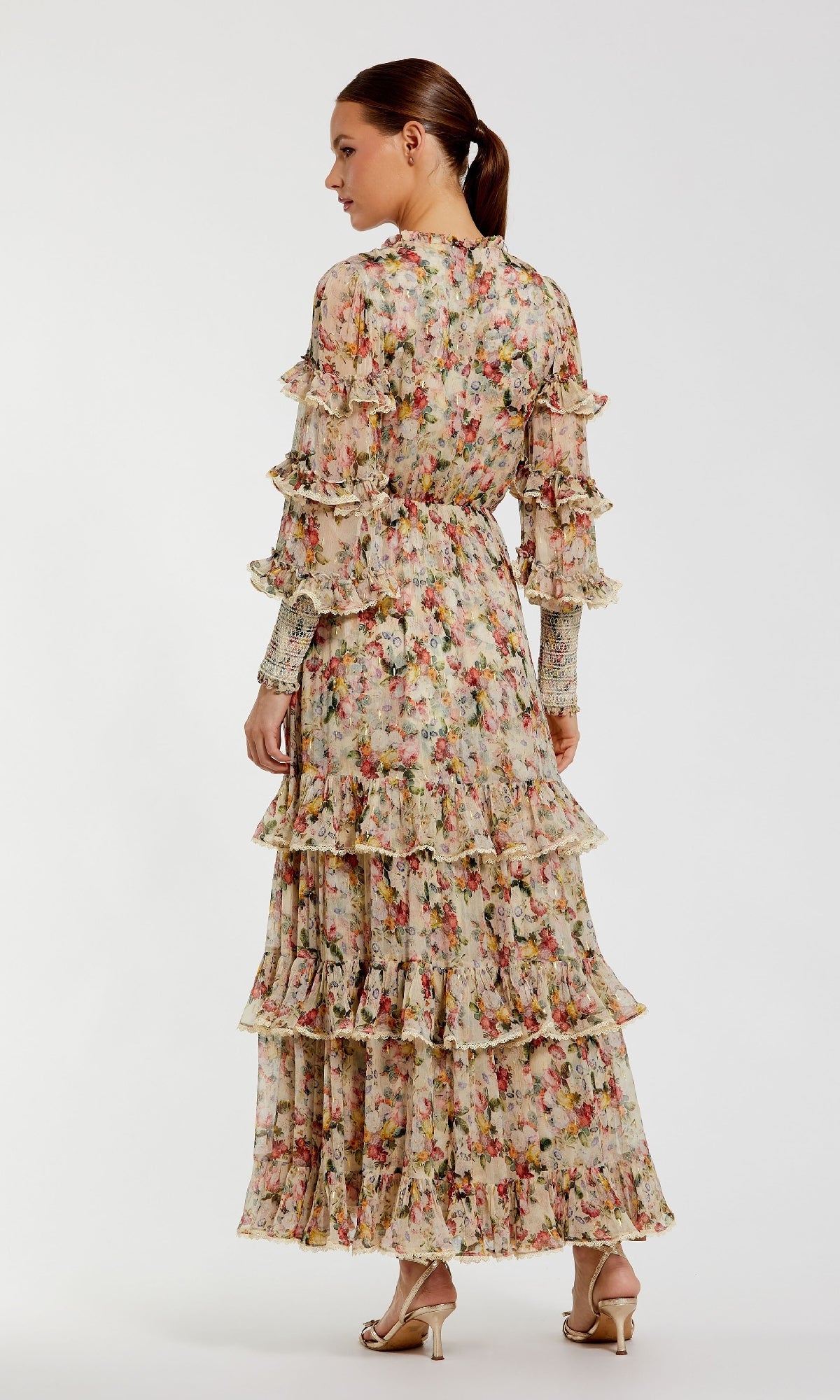 A woman stands facing away in the Mac Duggal 8334 Long Sleeve Ruffle Formal Dress, featuring a floral print, sheer sleeves, and tiered skirt; her hair is tied back and she's wearing heeled sandals against a plain background.