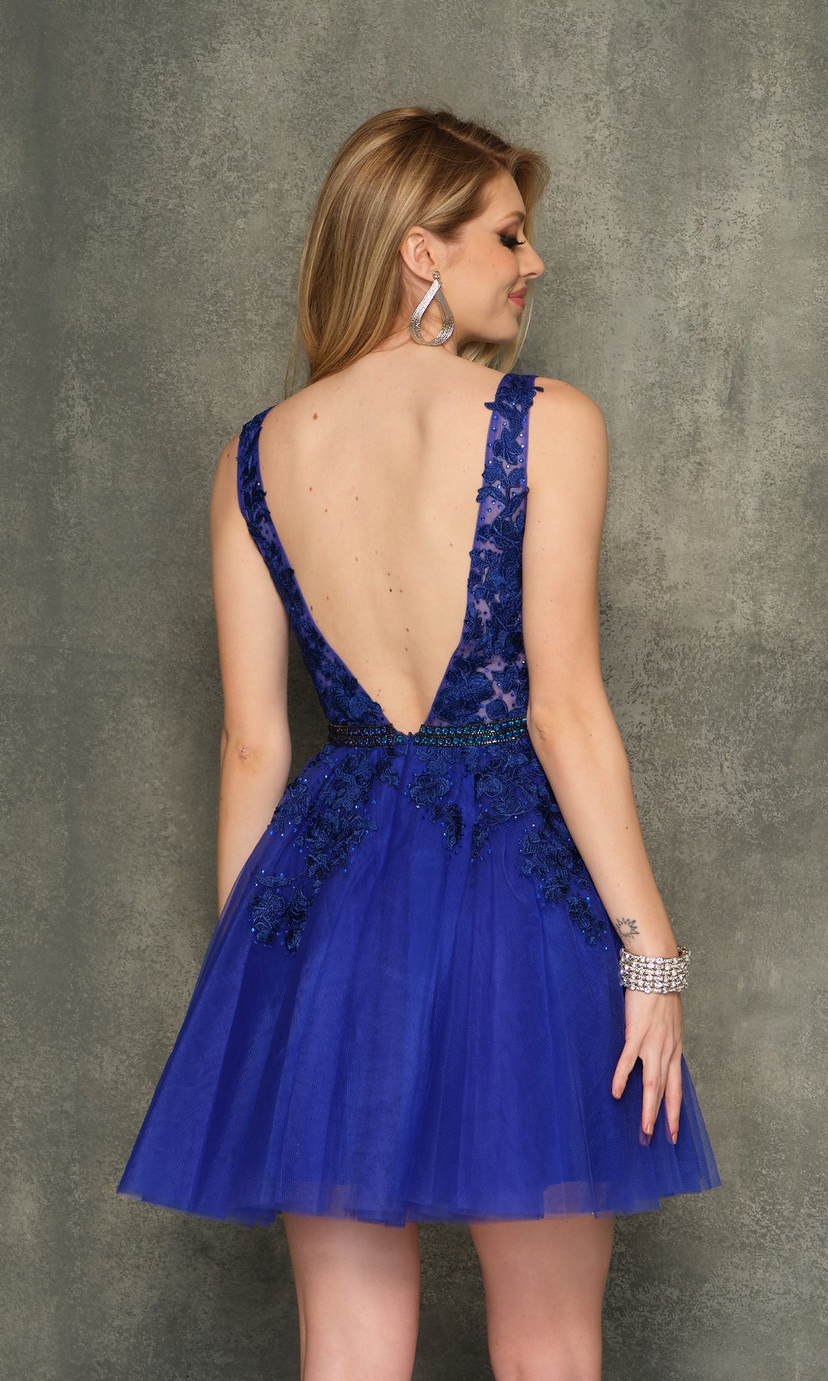 A woman wearing the A-Line Short Homecoming Dress H341—sleeveless, royal blue, with a deep V-back, lace and sequin details, and a beaded bodice—stands against a textured gray background.