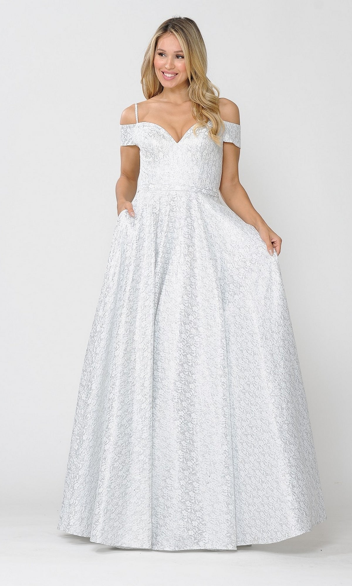 A woman smiles while posing in the Cold-Shoulder Silver and White Long Prom Gown 8380, an off white and silver evening dress, against a plain background, holding the sides of her gown.