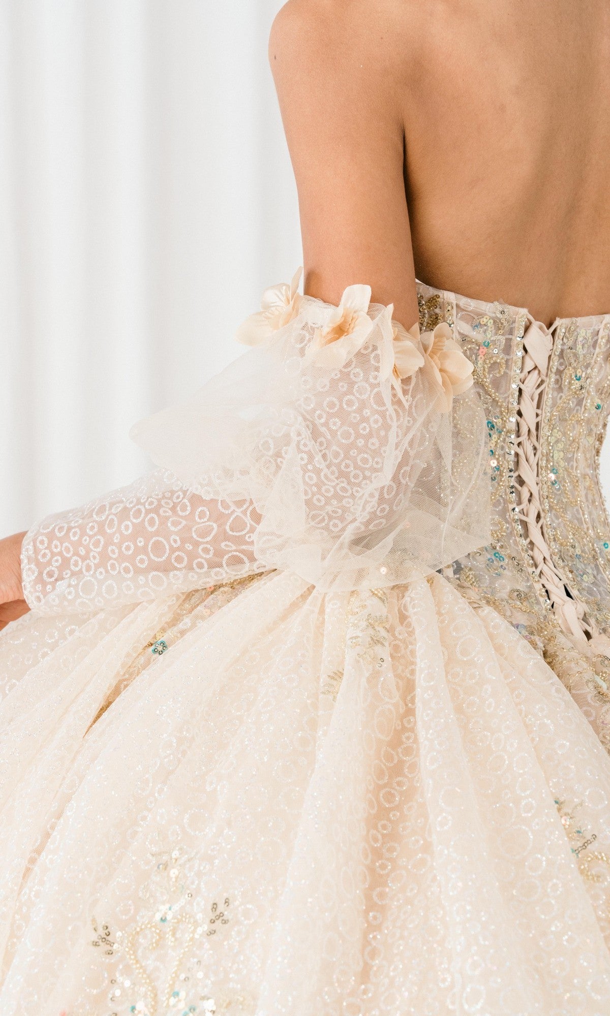 A close-up of a person's back wearing the Quinceanera Dress 8385J By Cinderella Couture, featuring an off-the-shoulder lace-up design, corset bodice, sheer puff sleeves, and intricate sequin embellishments.