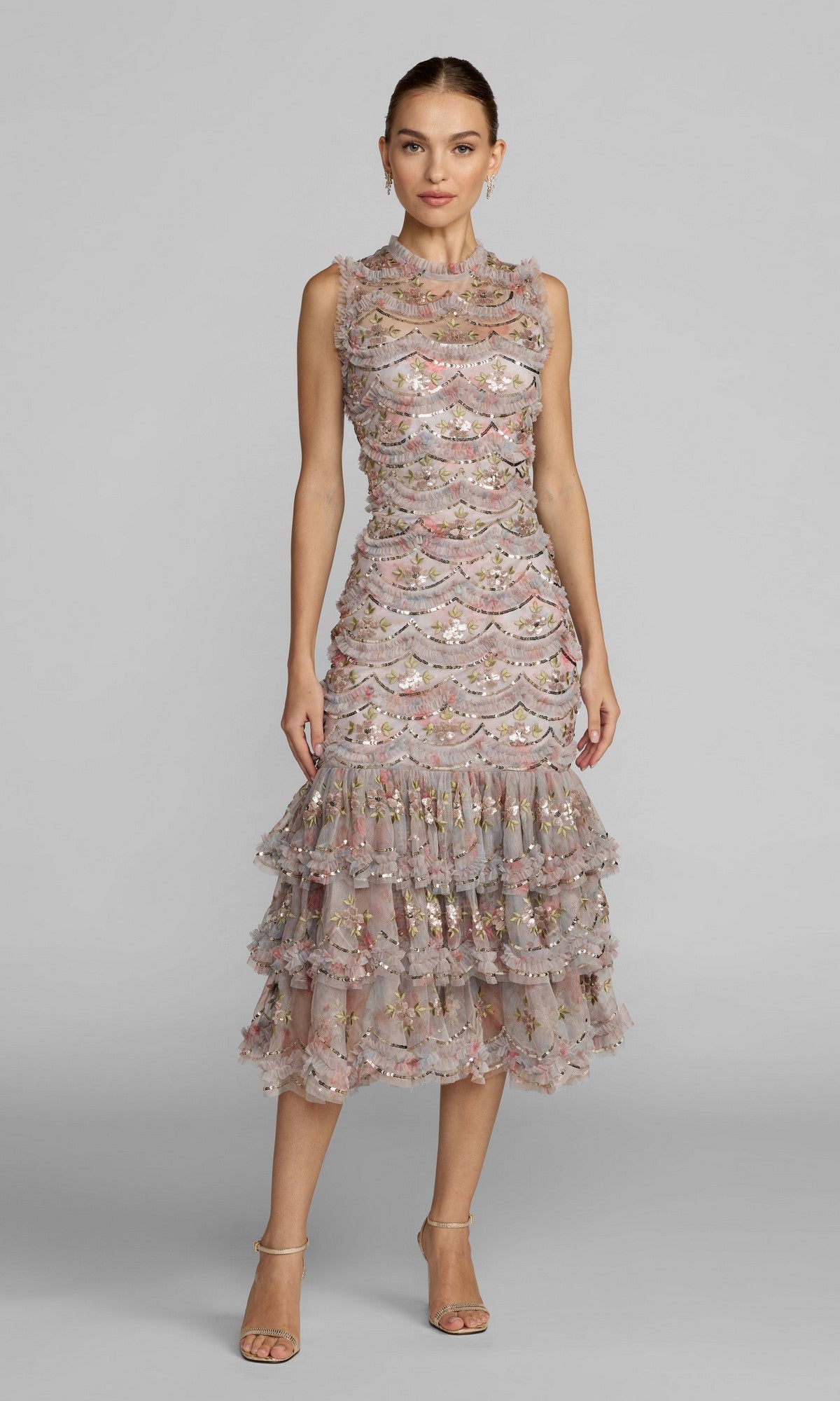 A woman stands against a plain gray background wearing the Midi Party Dress: Mac Duggal 8412 in Pastel Multi, a sleeveless floral dress with a high neckline and knee-length, tiered ruffle skirt, paired with open-toe heels.