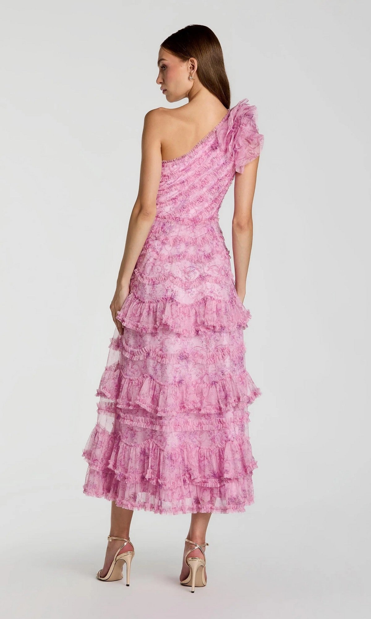 A woman faces away, wearing the Tea-Length Party Dress: Mac Duggal 8422 in pinkโfeaturing a one-shoulder design and ruffled, tiered midi skirt paired with strappy high heelsโstanding against a plain light background for a strikingly elegant look.