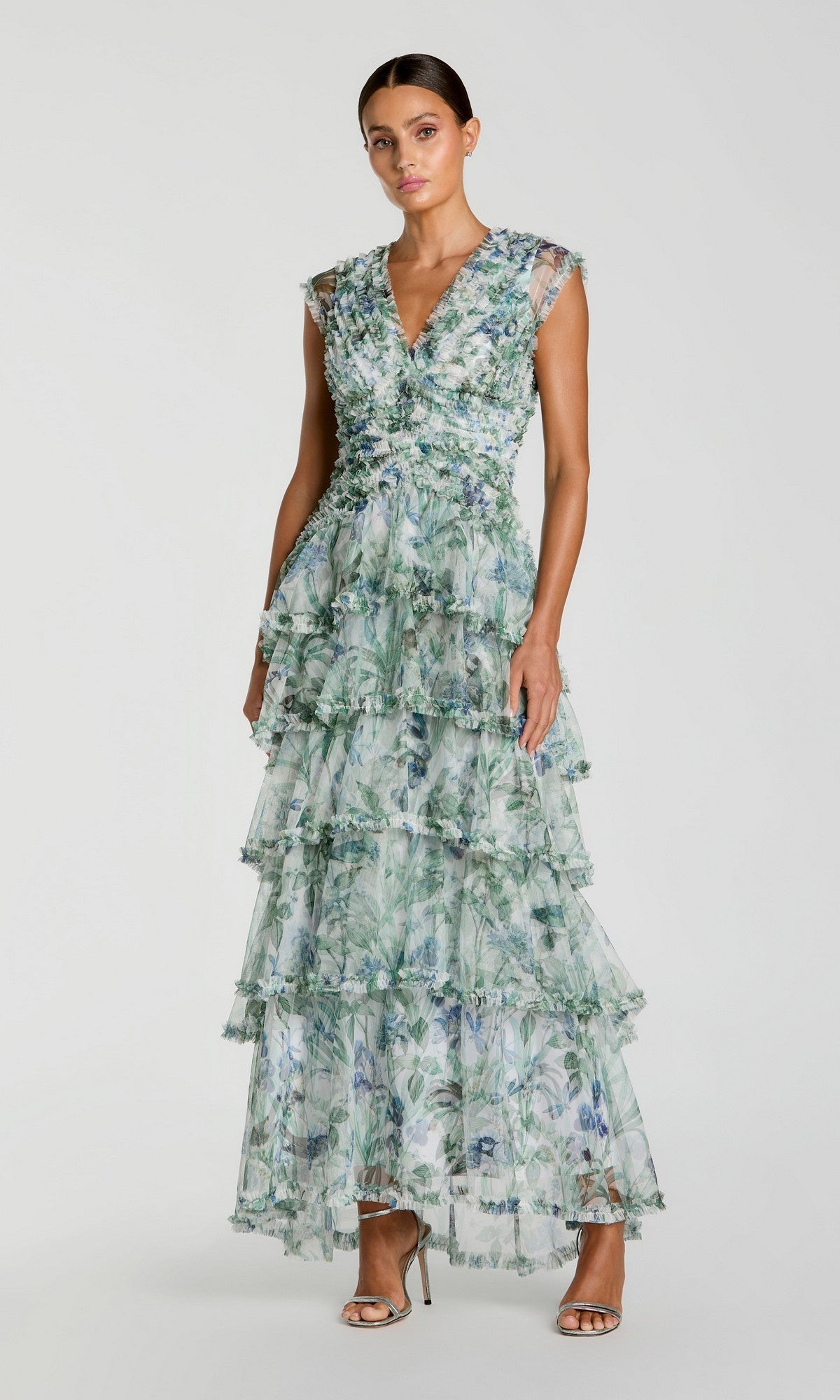 Woman wearing the Mac Duggal 8423 Long Formal Dress in a sleeveless, floor-length, ruffle A-line silhouette with a blue multi floral print, standing in heels against a plain background.