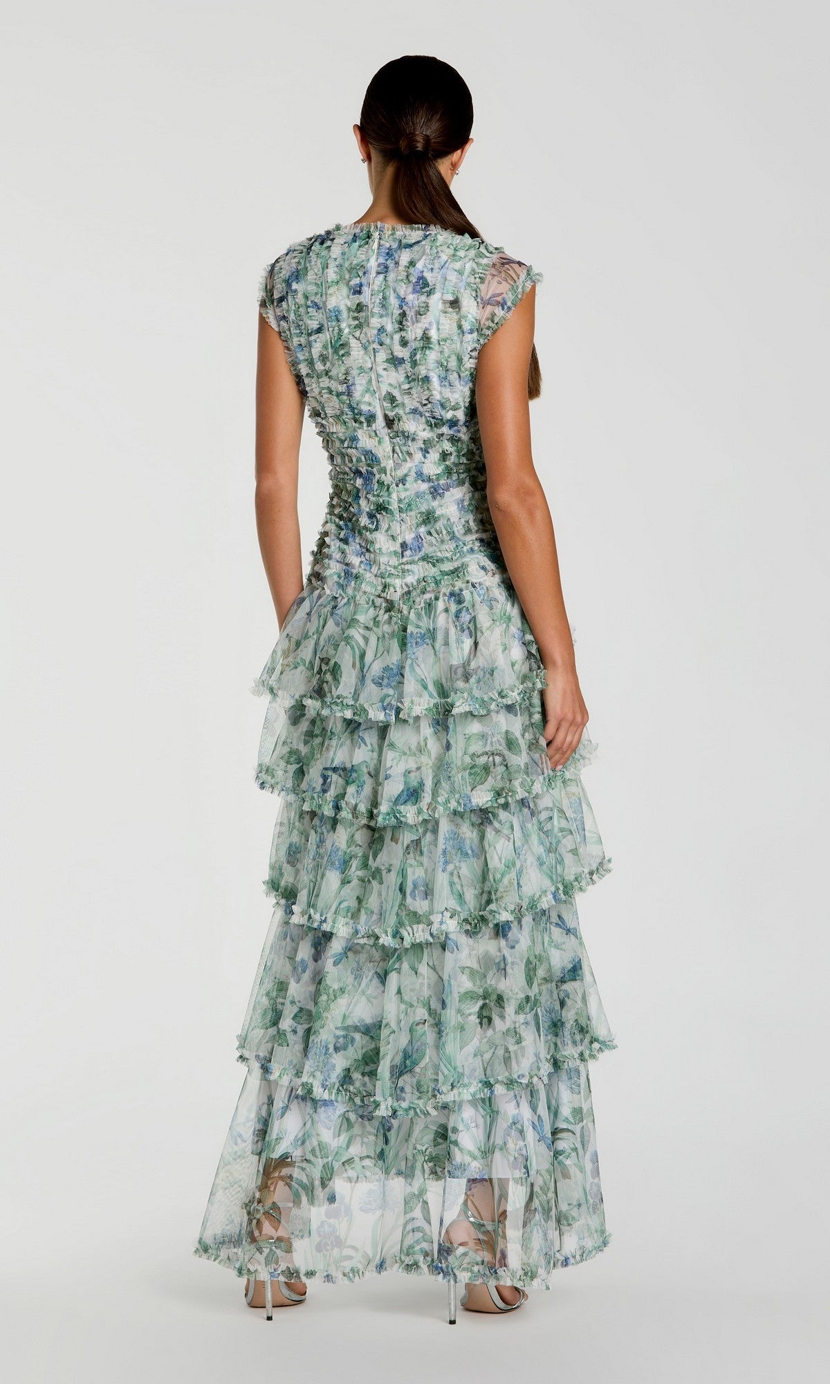 A woman stands facing away, wearing the Long Formal Dress: Mac Duggal 8423—a sleeveless, floor-length A-line gown featuring ruffles and a blue and green floral pattern—paired with high heels against a plain white background.