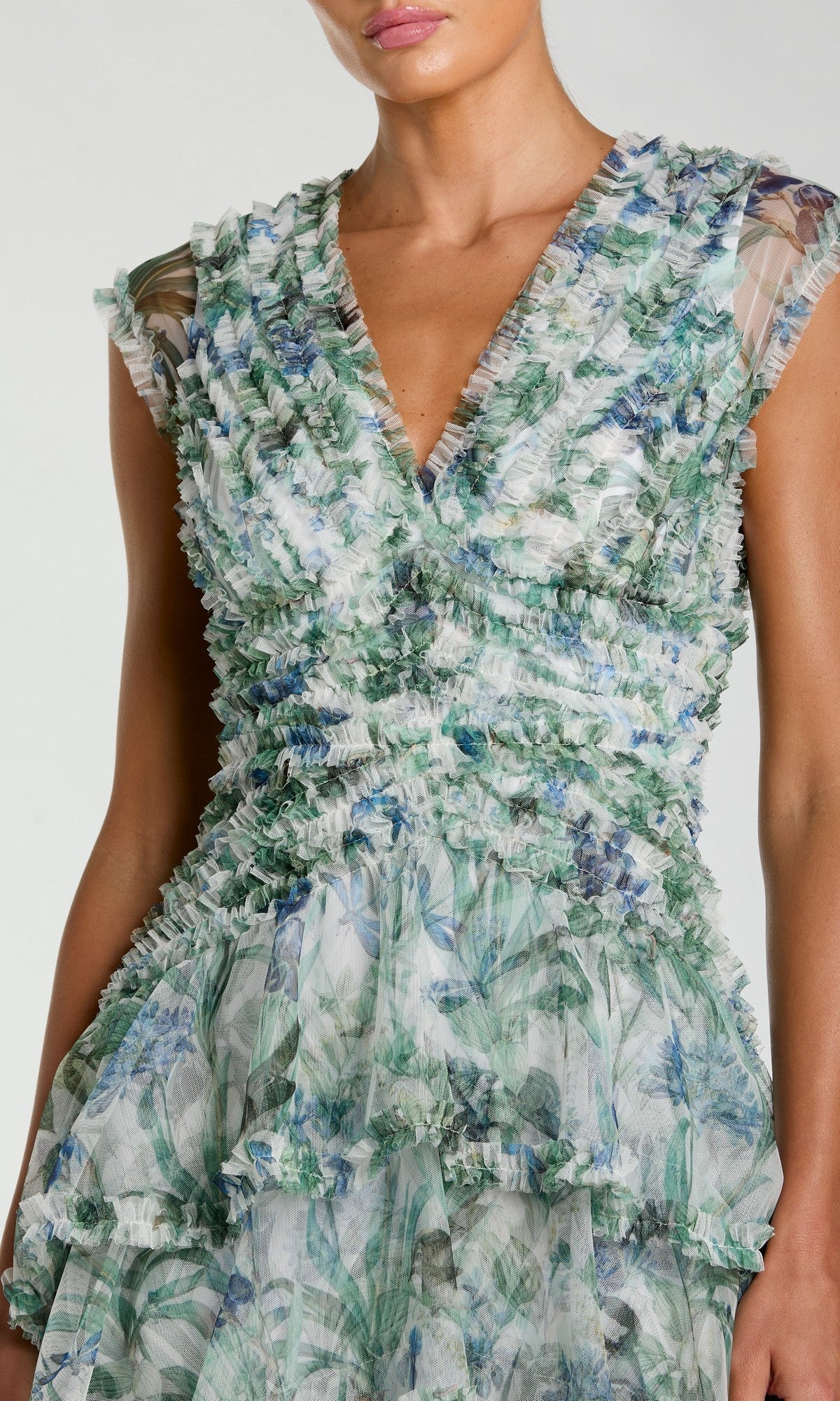 A woman wearing the Mac Duggal 8423 Long Formal Dress, featuring a sleeveless V-neck design with a blue floral pattern in green tones and ruffled detailing throughout the bodice and skirt.