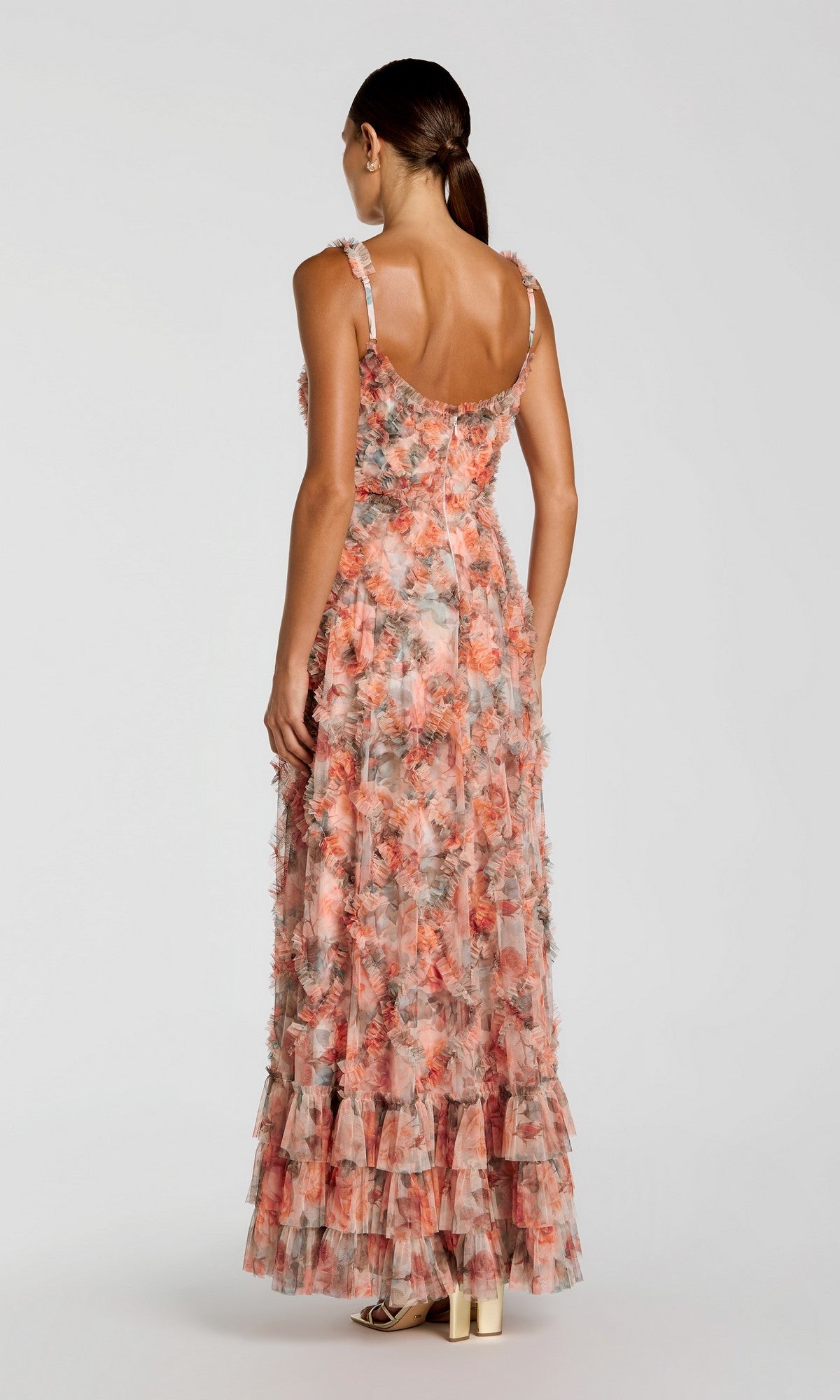 A woman stands with her back to the camera, wearing the sleeveless, floor-length Mac Duggal 8424 long formal dress featuring floral patterns, ruffled layers, and paired with high heels.