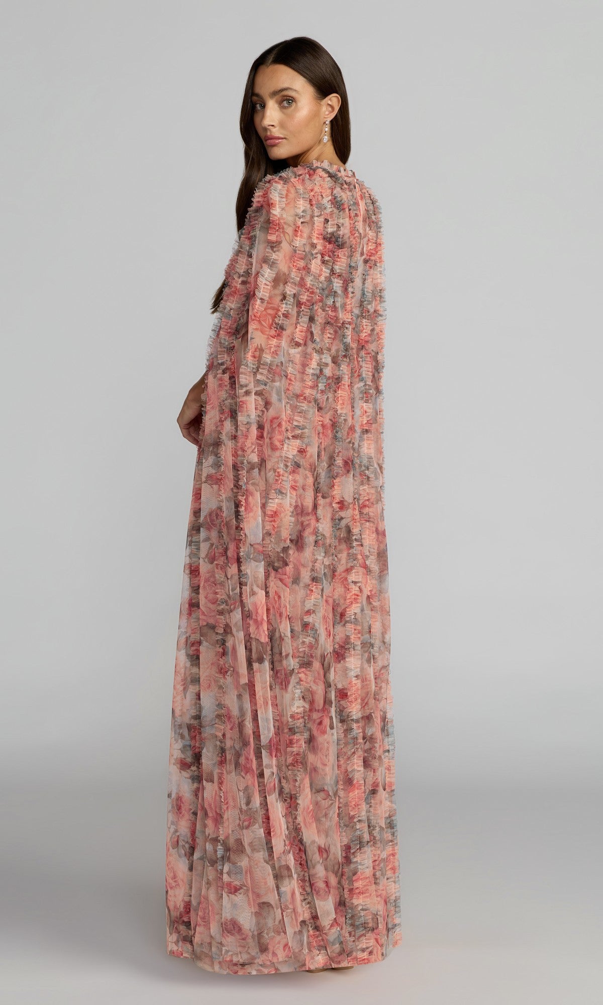 A woman stands looking over her shoulder, wearing the Mac Duggal 8431 long formal dress—a flowy, pink floral gown crafted from sheer fabric—set against a neutral background.