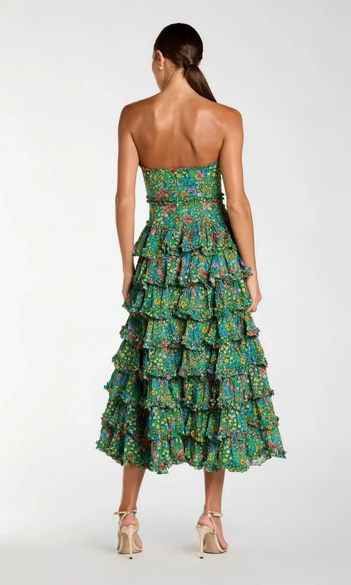 A woman, her hair tied back in a low ponytail, faces away in the Mac Duggal 8459 Tea-Length Party Dress—a strapless green floral-printed design with tiered ruffles—paired with beige high-heeled sandals, set against a plain light background.