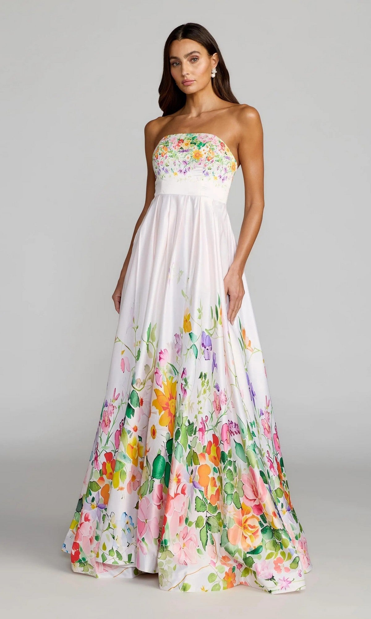Woman wearing the Mac Duggal 8470 long formal dress, a strapless satin gown adorned with a vibrant floral print on the bodice and skirt, standing against a plain gray background. This floor-length design brings an elegant and modern touch to classic eveningwear.