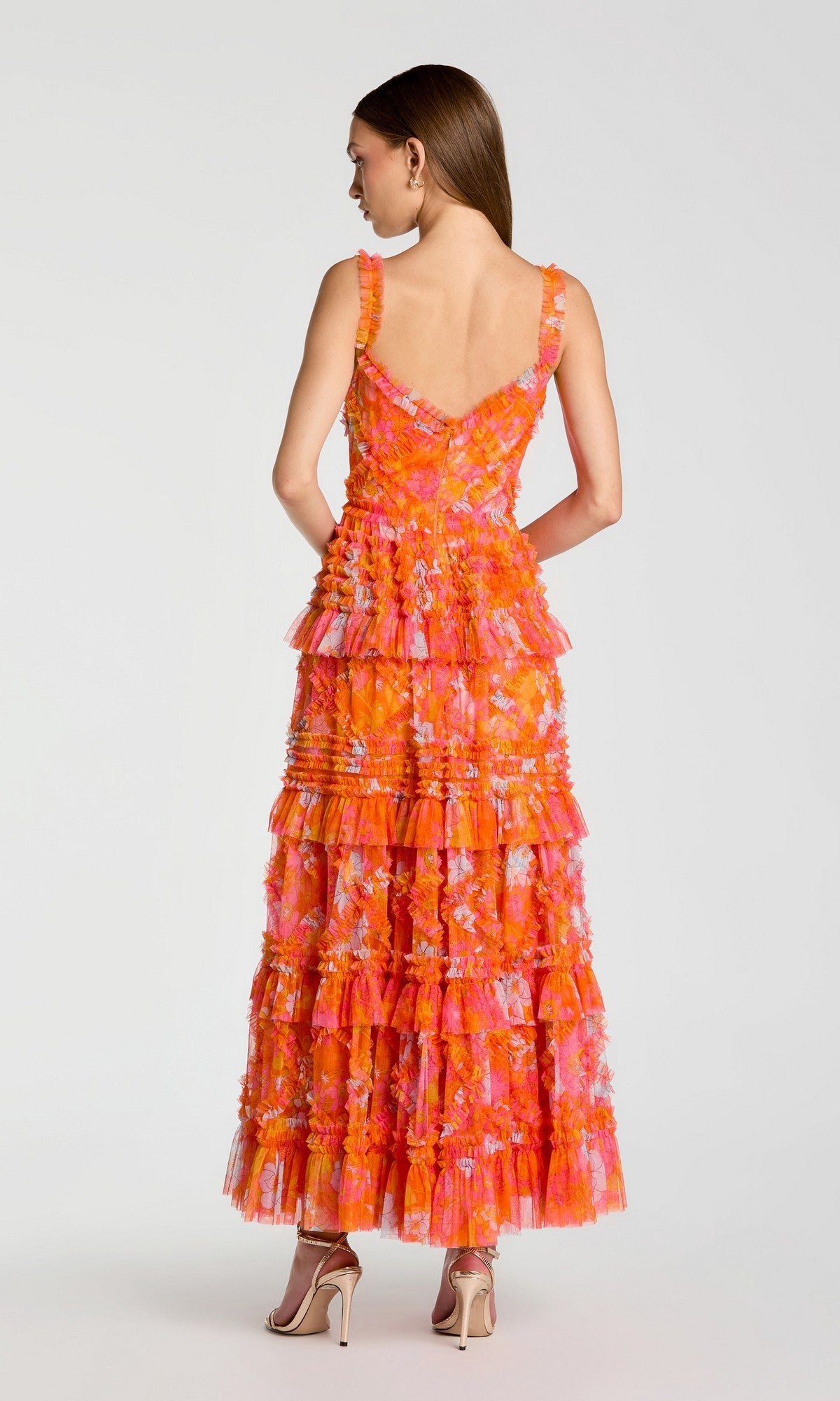 A woman stands facing away, wearing the Mac Duggal 8475 long formal dress— a sleeveless, floor-length, layered fit-and-flare gown in orange and pink with floral details and ruffles—paired with beige high-heeled sandals.