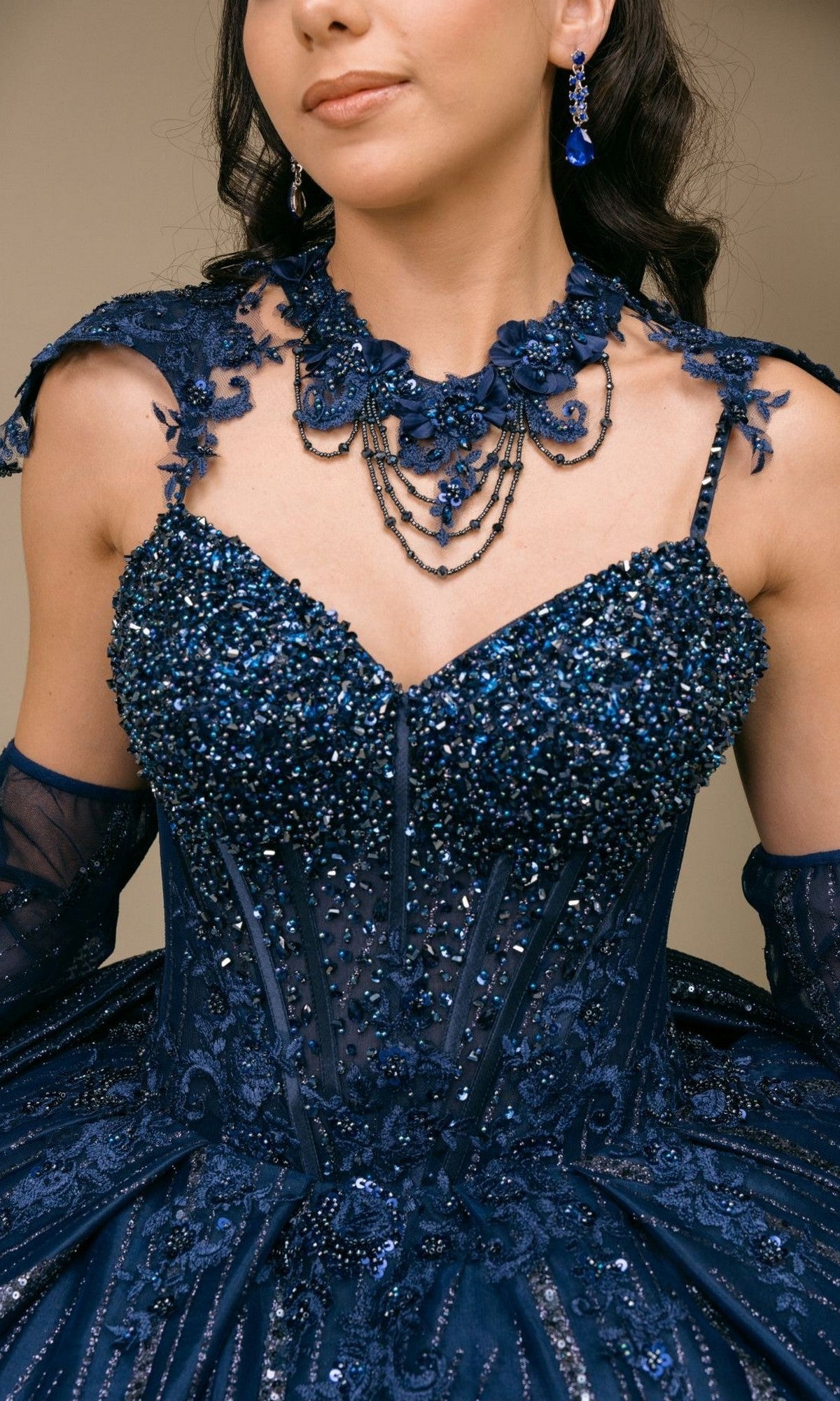 A woman is wearing the Quinceanera Dress 8510JQ By Cinderella Couture—a dark blue sequin-embellished gown with sheer sleeves—paired with a matching necklace and earrings, photographed from the shoulders to the waist.