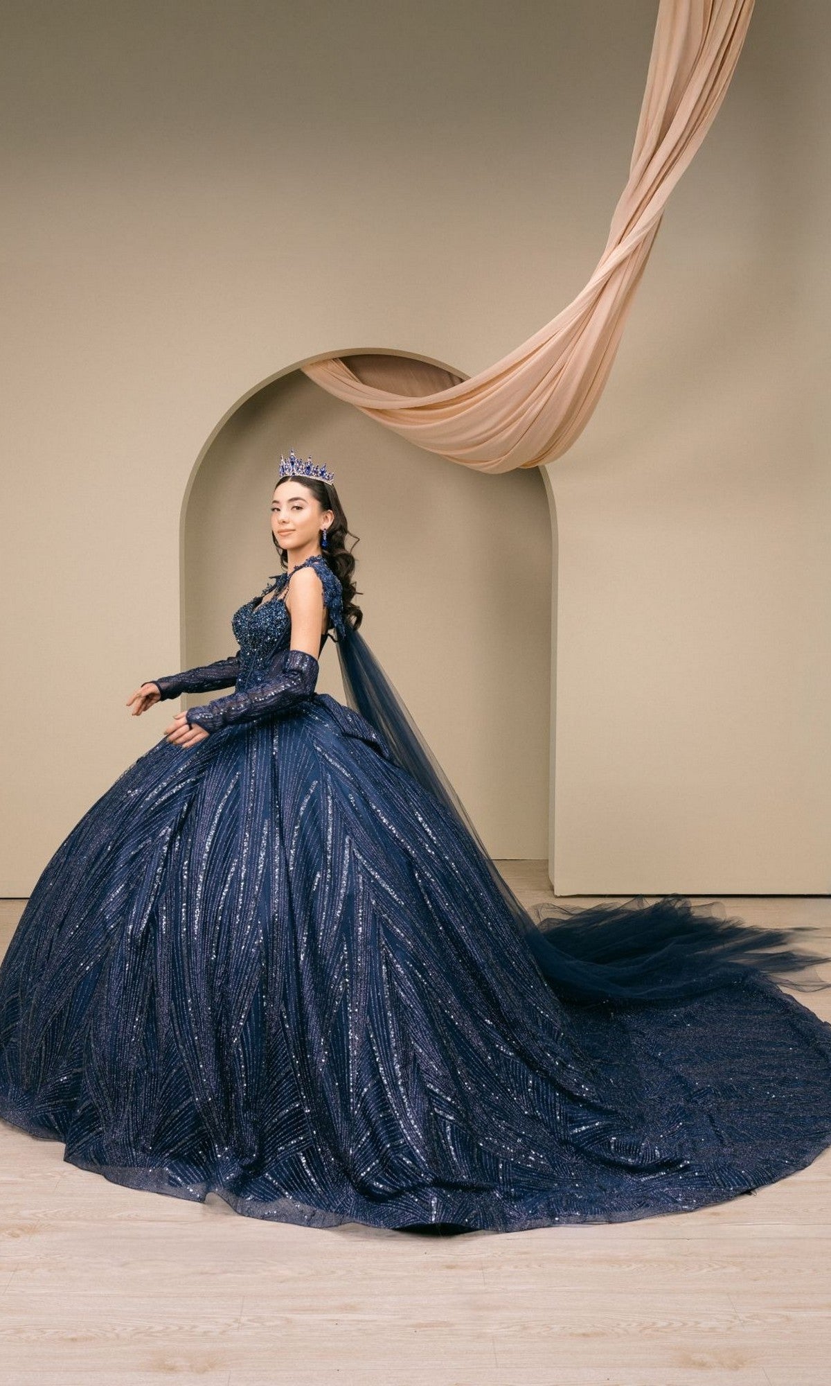 A woman wearing the Quinceanera Dress 8510JQ by Cinderella Couture—a dark blue, sequin-embellished ball gown—and a crown stands before an arched backdrop draped with fabric, radiating timeless elegance.