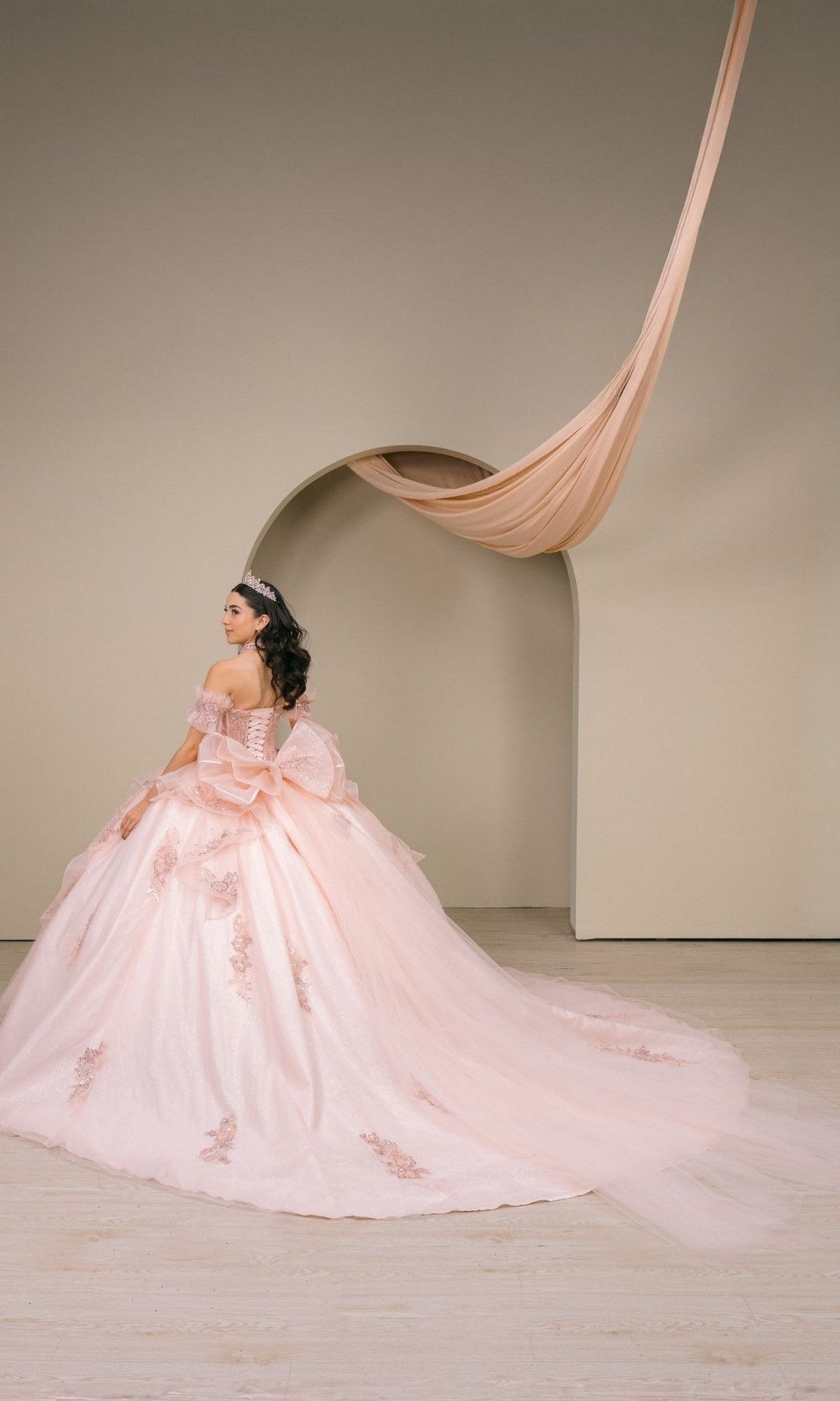 A woman sits gracefully in front of a beige wall, framed by an arch and elegantly draped fabric above, wearing the Quinceanera Dress 8540JQ by Cinderella Couture—a blush pink gown adorned with delicate floral details.