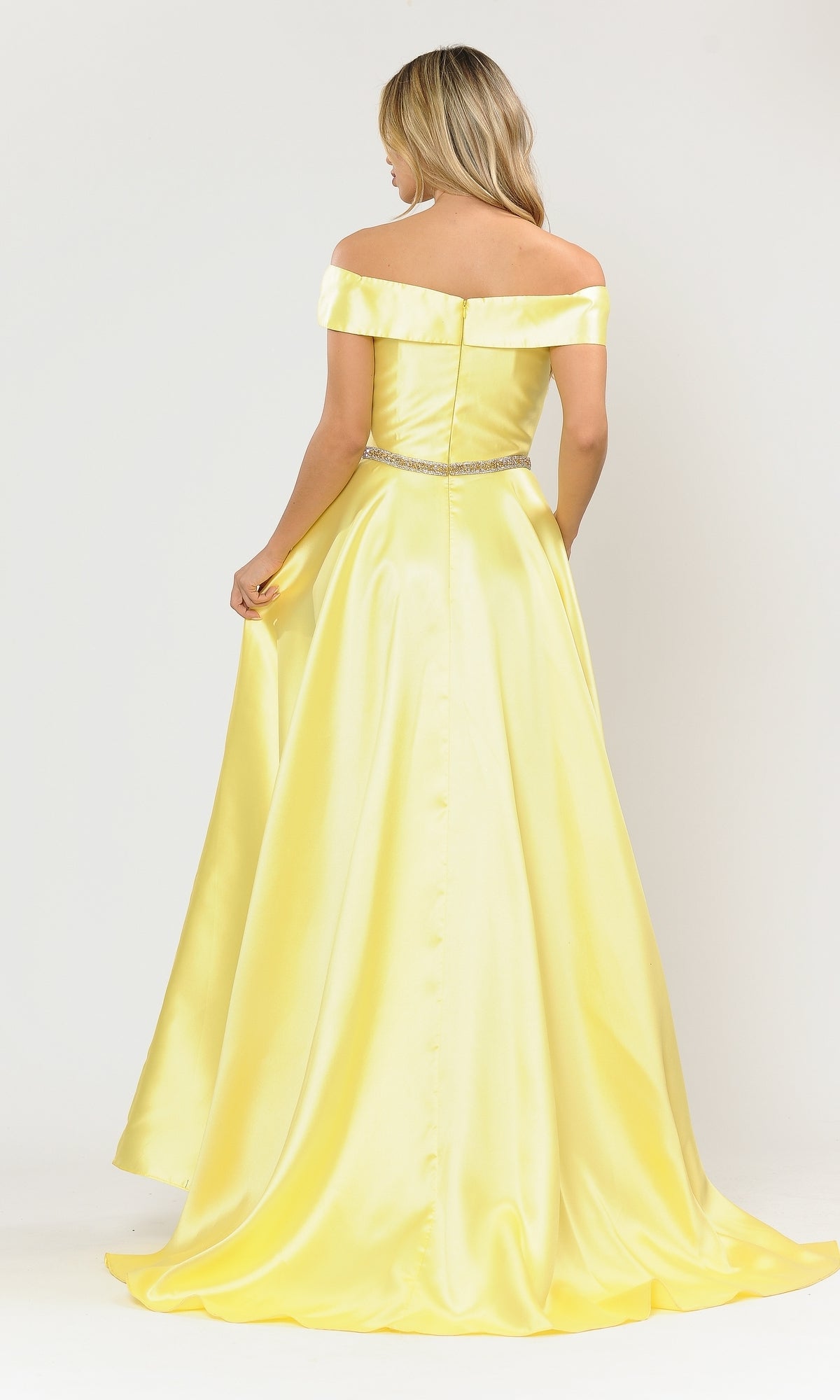 A woman in the Plunging V-Neck Long Mikado Prom Ball Gown 8680, featuring yellow satin and a jeweled waistband, stands facing away while holding her skirt out slightly.