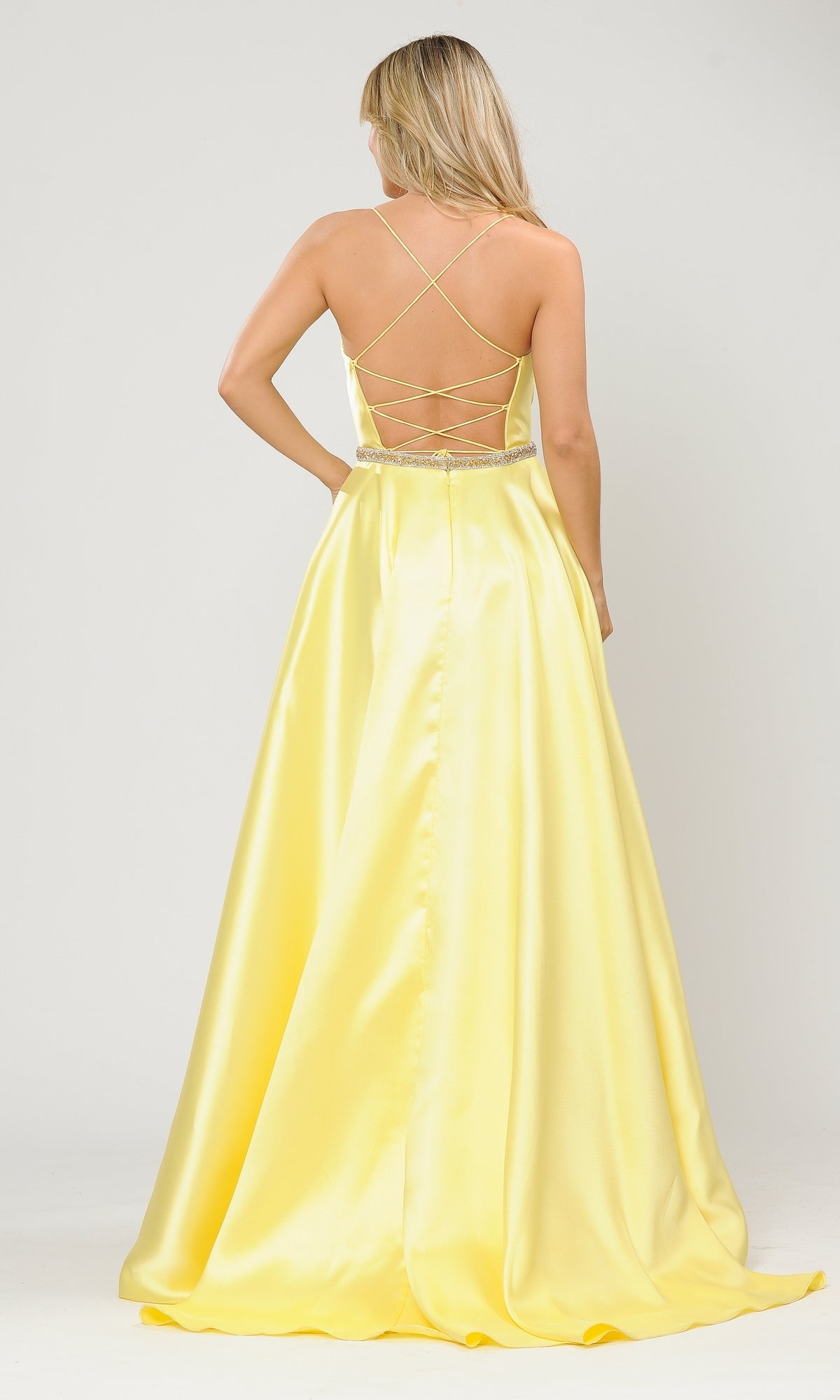 A woman in the Scoop-Neck Long A-Line Prom Dress with Pockets 8684, featuring yellow satin, a crisscross open back, jeweled waistband, and floor-length design, stands against a plain white background.