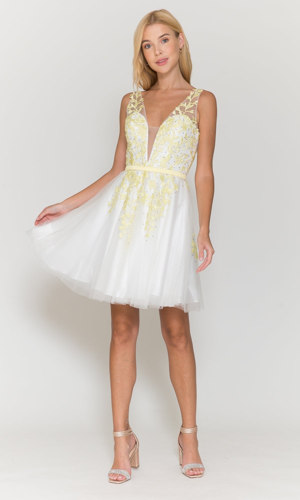 A woman wearing the Tulle and Lace Short A-Line Homecoming Dress 8696, featuring a sleeveless design, white fabric with yellow floral embroidery, deep V neckline, and sheer skirt stands against a plain white background.