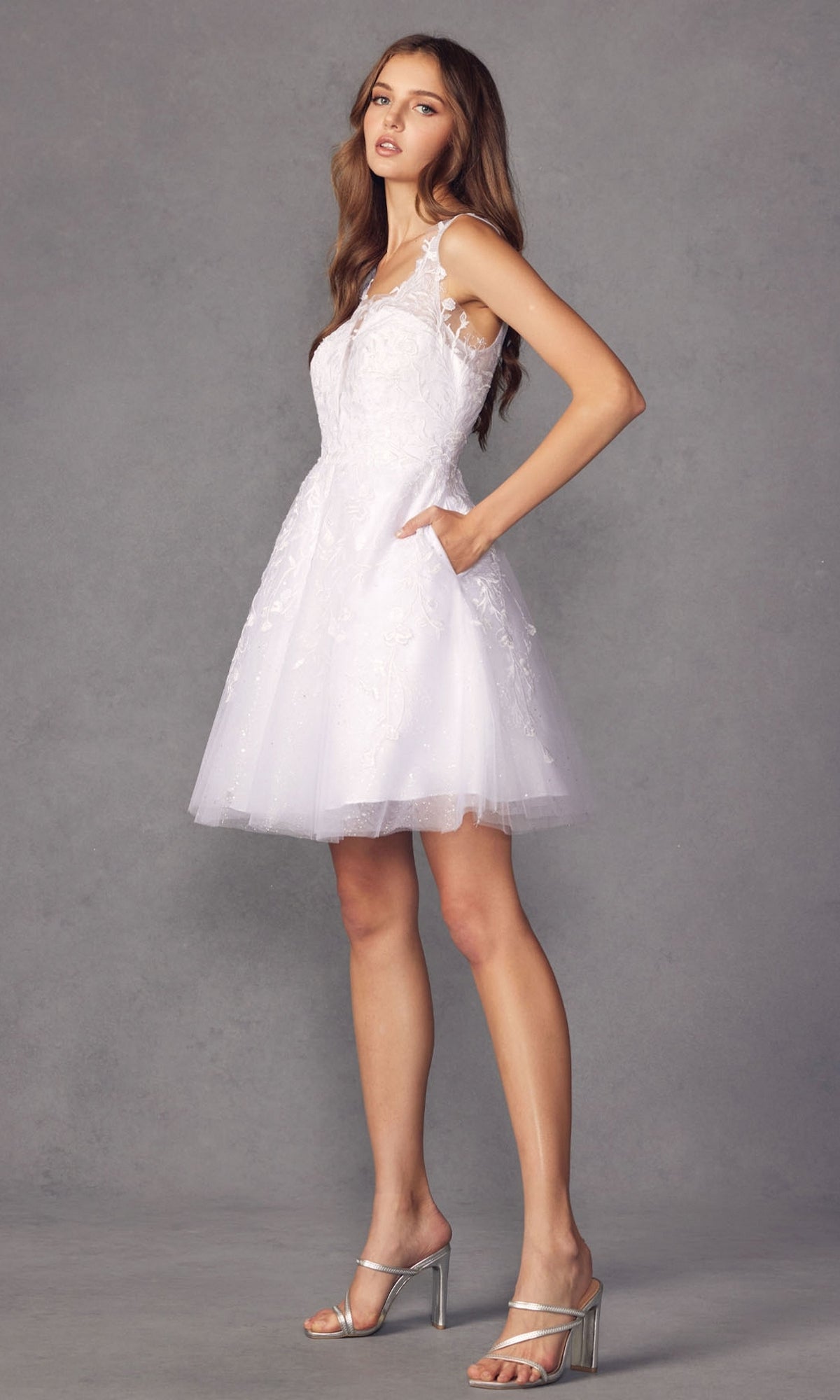 A woman poses against a plain gray background wearing the Short Glitter-Floral A-Line Homecoming Dress 871, a white sleeveless semi-formal party dress with lace details, paired with silver high-heeled sandals.