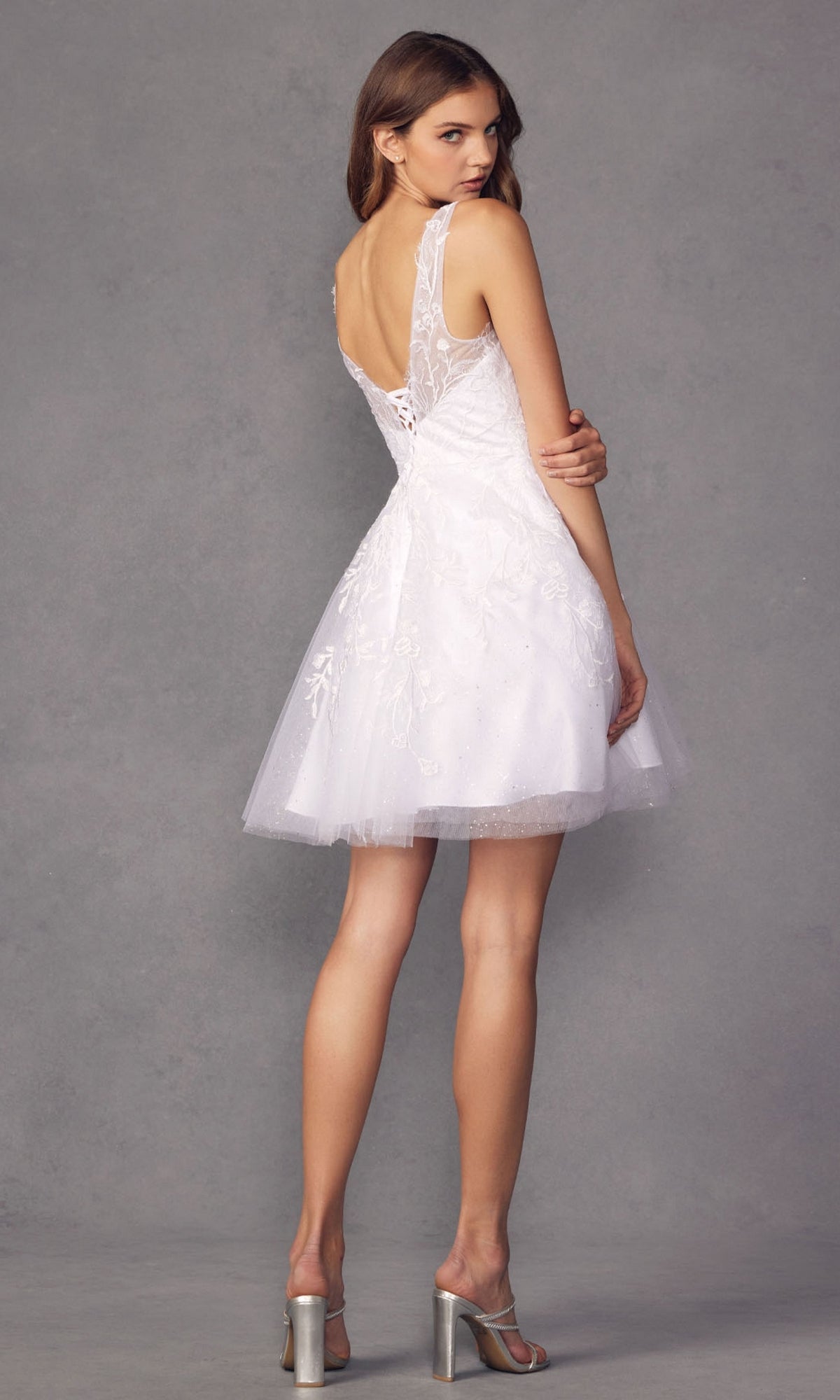 Woman in the Short Glitter-Floral A-Line Homecoming Dress 871, featuring a fitted bodice and flared skirt, stands in profile and looks over her shoulder against a plain gray background.