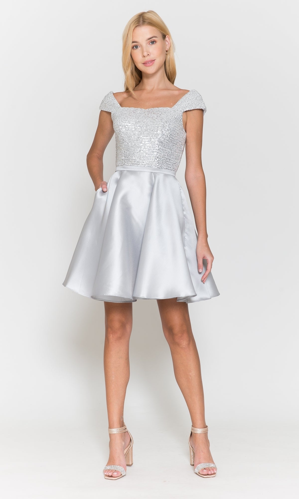 A woman stands wearing the Cap-Sleeve Short A-Line Homecoming Dress 8732, featuring a sequined bodice and off-the-shoulder sleeves, paired with strappy high-heeled sandals.