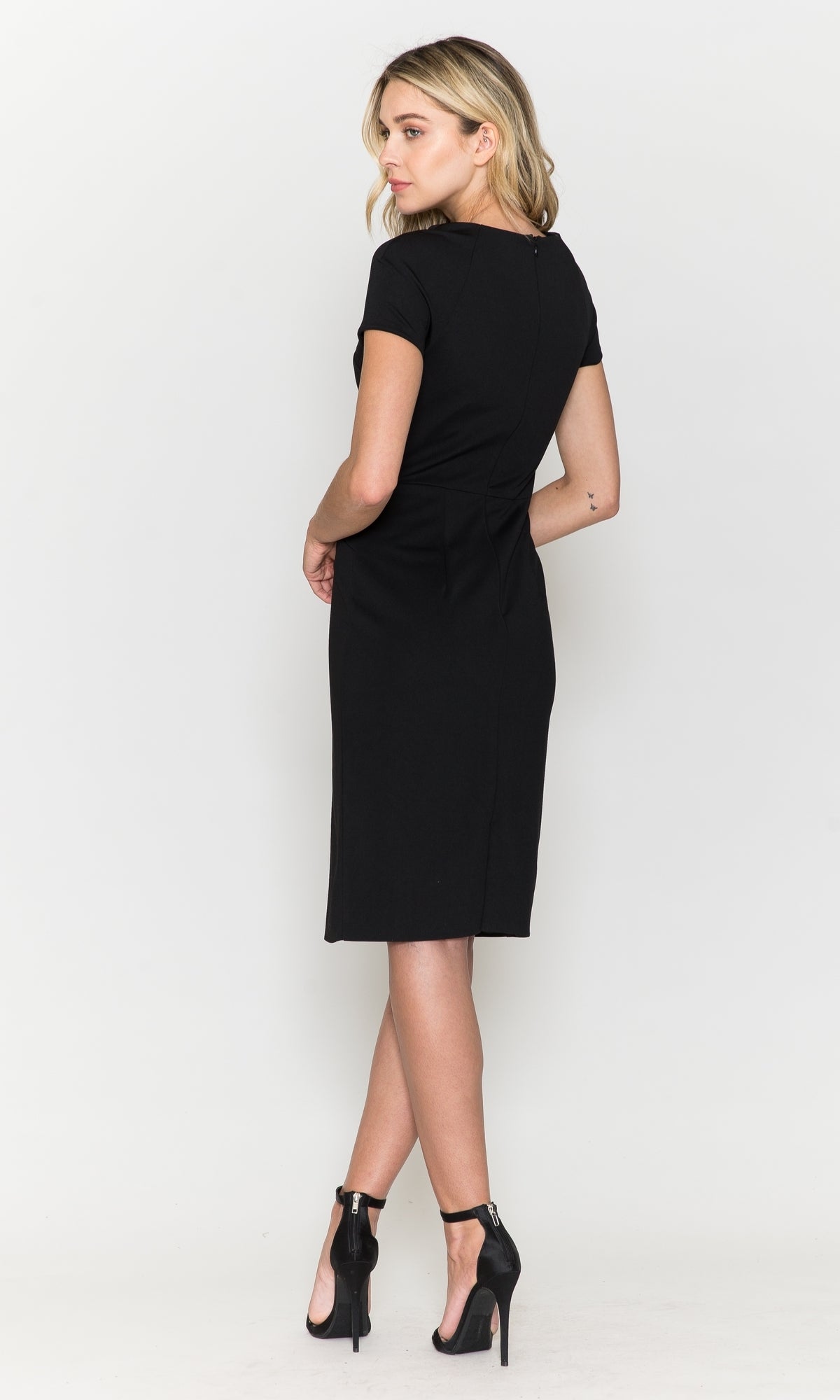 A woman faces slightly away, wearing the Short Sleeve Knee-Length Semi-Formal Dress 8774 in black with high heels, set against a plain white background.