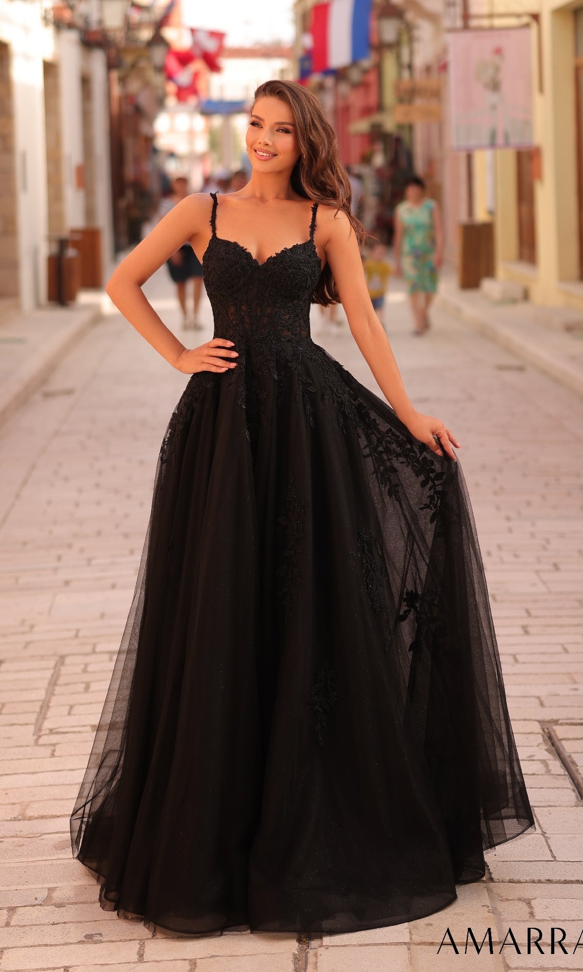 A woman stands on a cobblestone street wearing the Long Formal Dress 88109 by Amarra, a black lace embroidered evening gown with spaghetti straps.