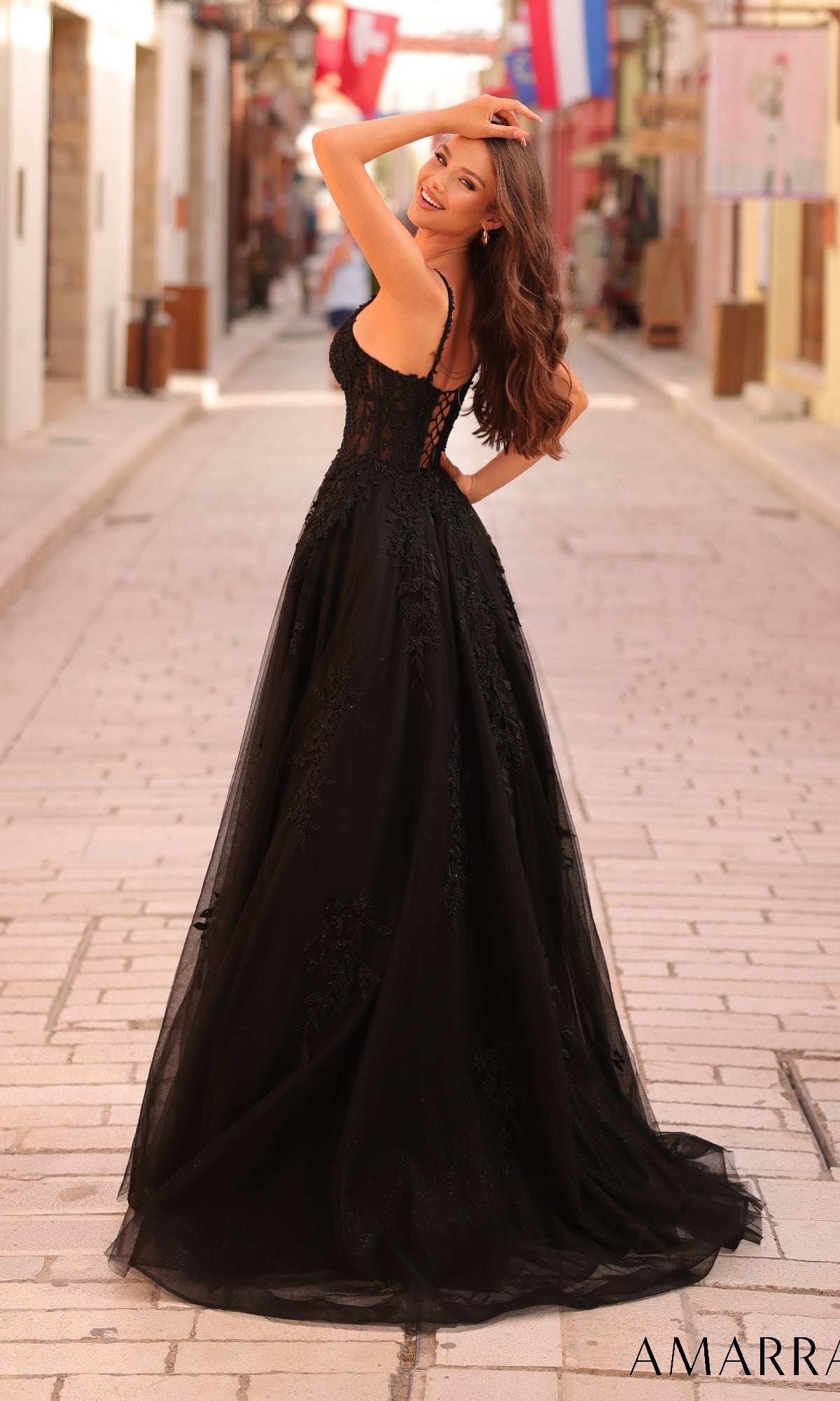 Wearing the Long Formal Dress 88109 by Amarra, a woman in a black lace-embroidered gown with a sheer corset bodice stands on a cobblestone street, looking over her shoulder and smiling.