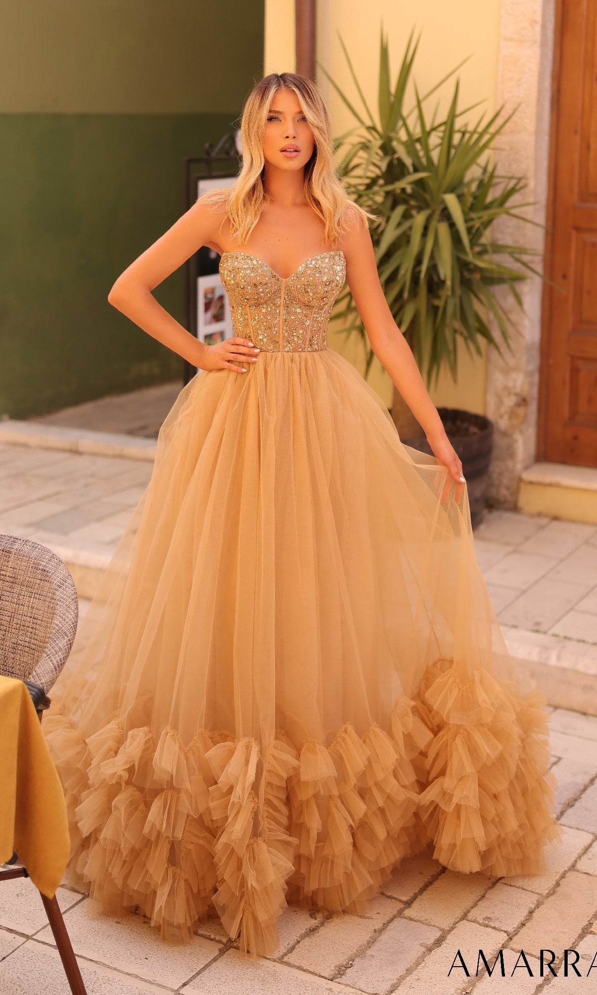 A woman stands outdoors wearing the Amarra 88339 long prom dress, which features a strapless sequined corset bodice and a gold tulle floor-length skirt with a ruffled hem.