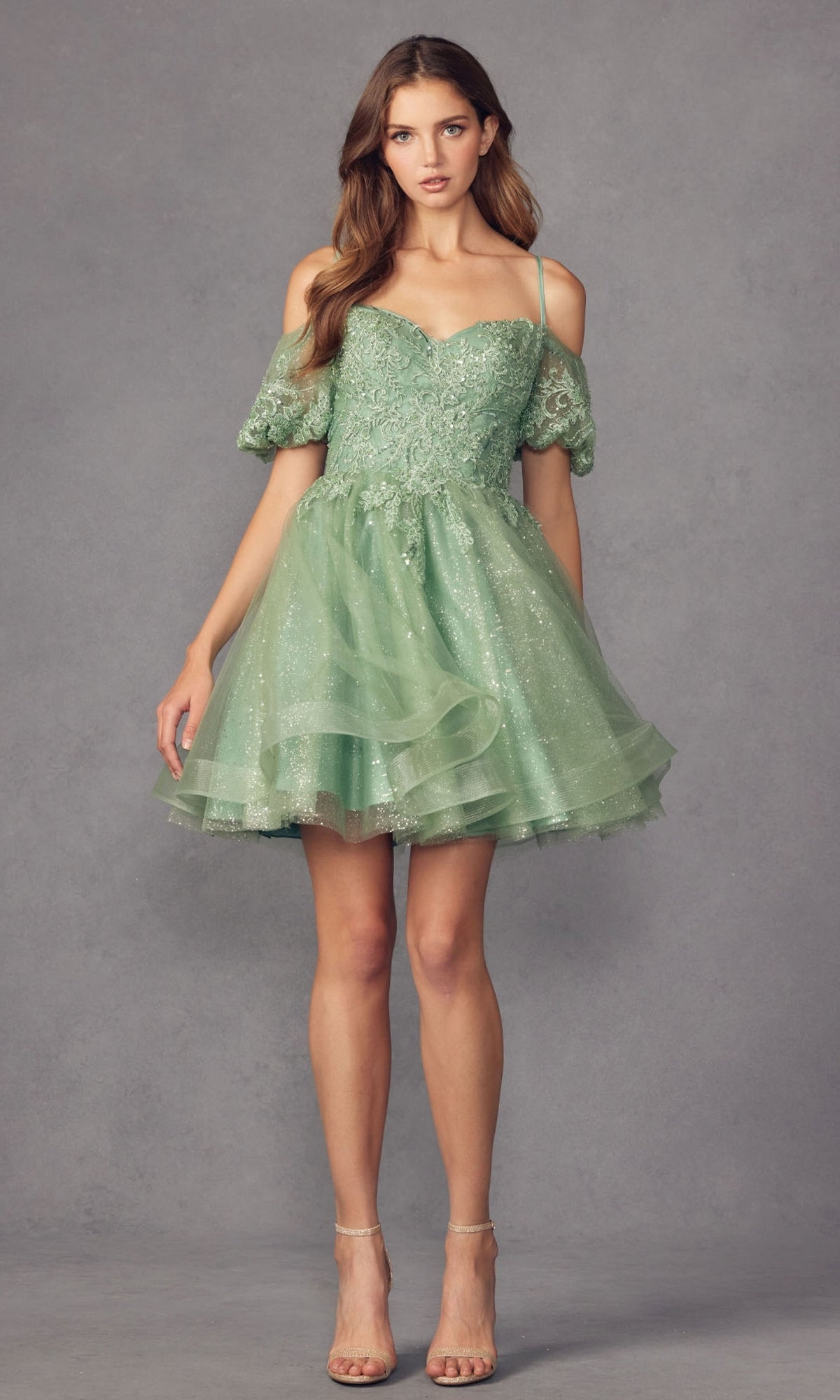 Against a gray background, a woman wears the affordable Puff-Sleeve Short A-Line Shimmer Prom Dress 886—a light green, off-shoulder style with lace and tulle accents—paired with strappy high-heeled sandals.