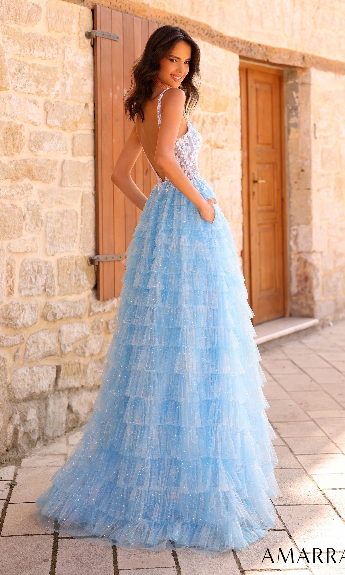A woman in the Amarra Deep V-Back Long Ruffled Prom Dress 88833 stands outdoors by a stone wall and wooden door, looking over her shoulder.