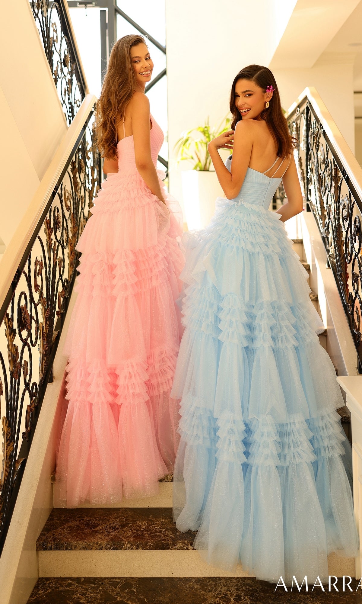Two women stand on a staircase, smiling over their shoulders; one wears the Amarra One-Shoulder Long Ruffled Prom Gown 88863 in pink, while the other wears a similar blue ruffled gown.