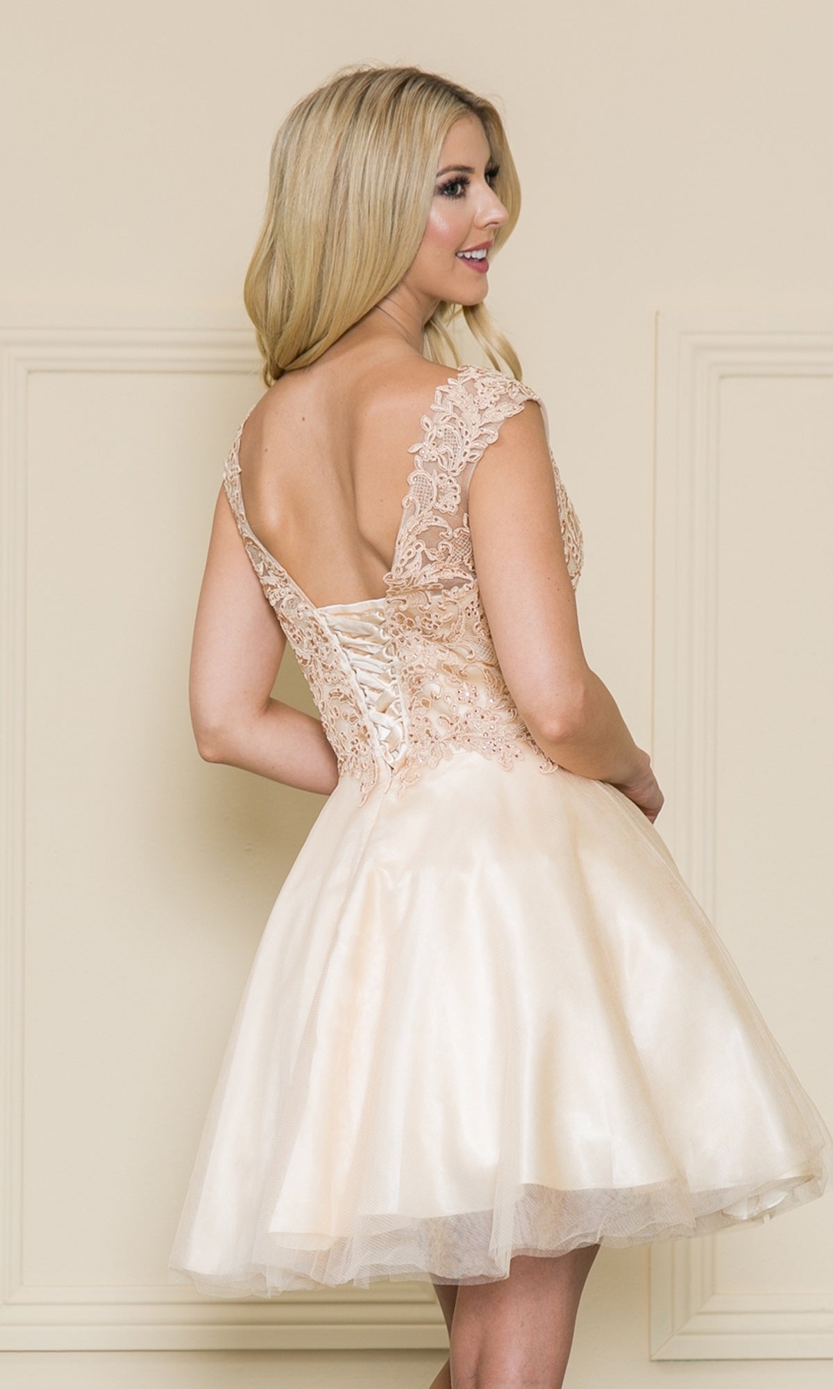 Smiling woman in the Lace-Bodice Short Babydoll Homecoming Dress 8902—a light beige A-line party dress with a lace-up back and delicate lace bodice—stands facing slightly away from the camera.
