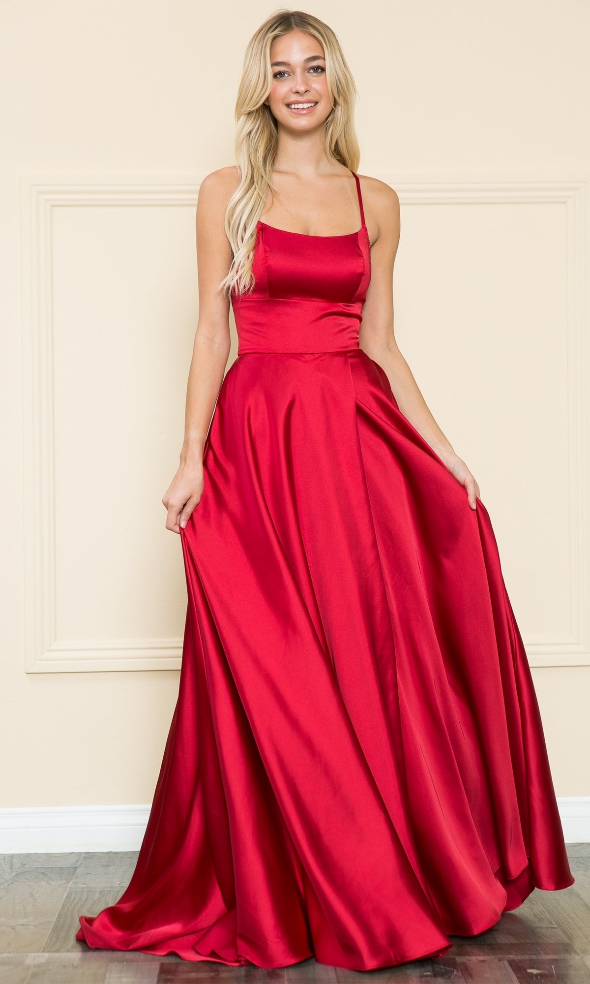 A woman stands in front of a beige wall wearing the Lace-Up Corset-Back Long A-Line Prom Dress 8910, featuring a floor-length, sleeveless red satin design with a fitted bodice and flared skirt.