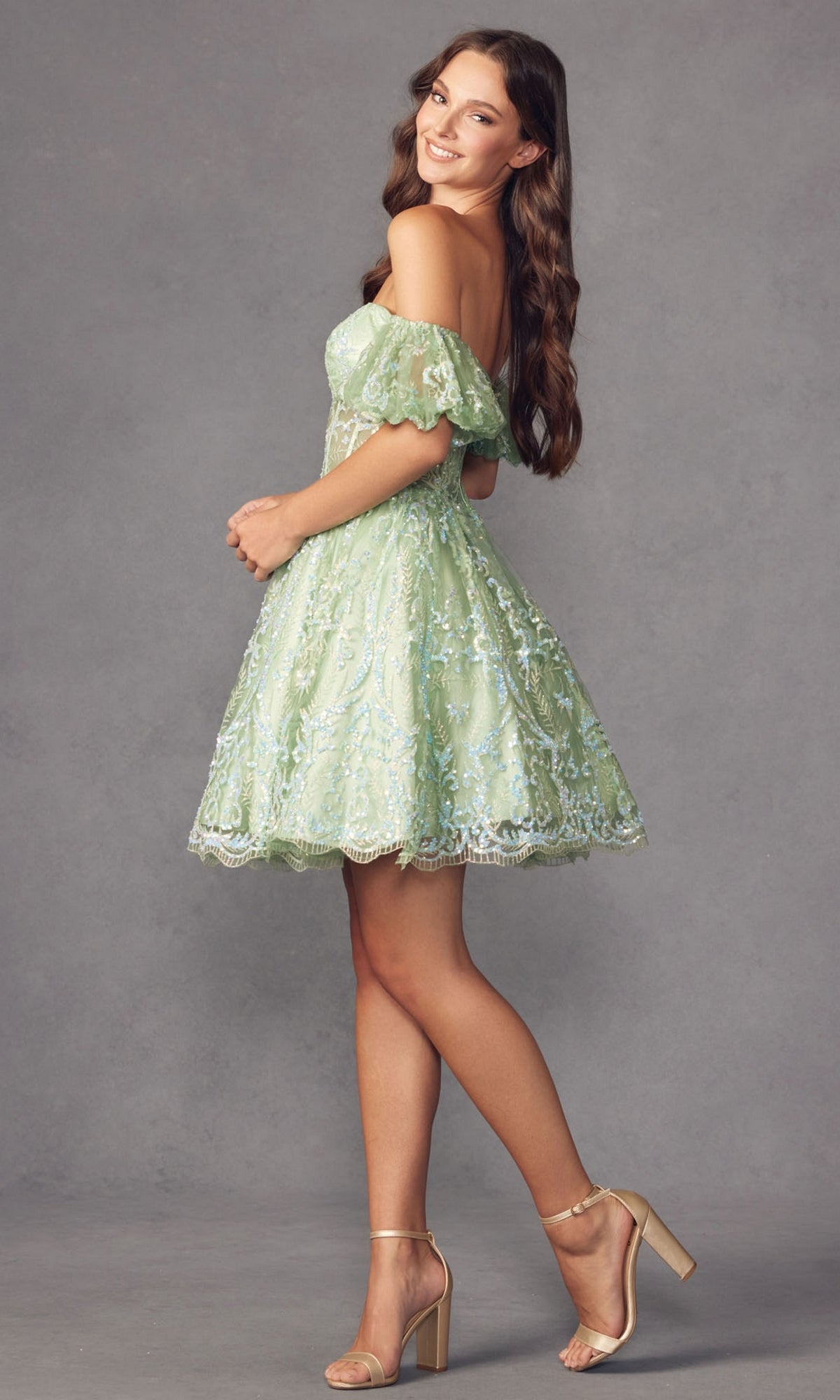 A young woman wears the Off-Shoulder Sheer-Corset Short Prom Dress 891, a light green lace dress with puff sleeves and a corset waist, paired with beige heels, as she poses against a plain gray background.