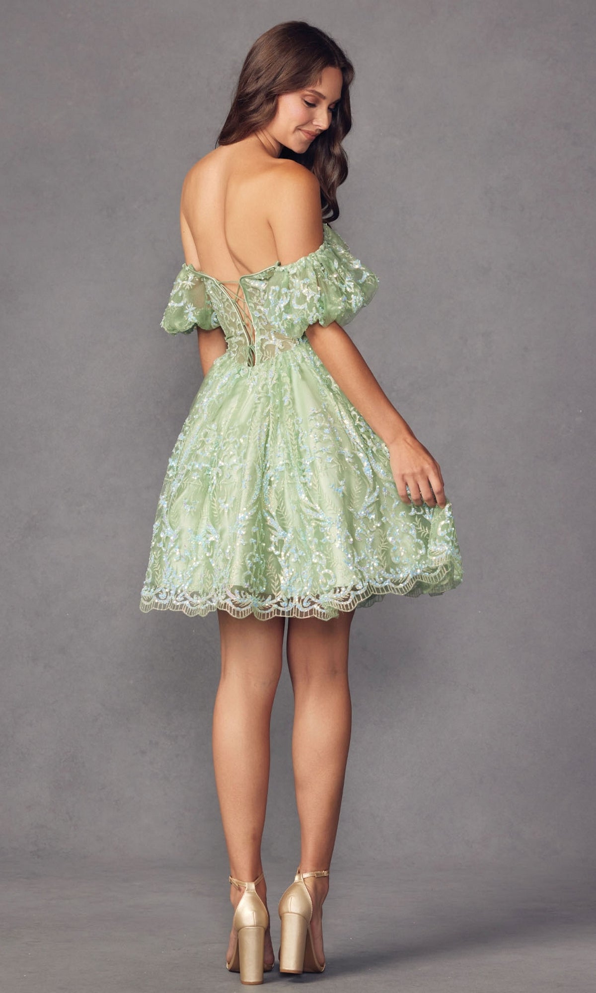 A woman in the Off-Shoulder Sheer-Corset Short Prom Dress 891—light green with sequins and a corset waist—paired with gold heels, stands against a gray background, facing slightly away.